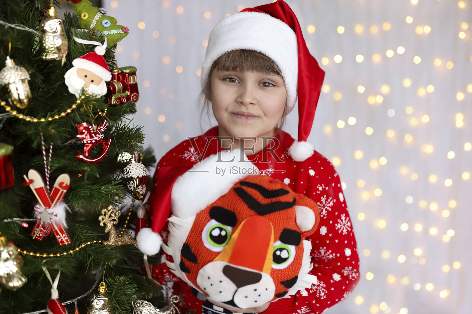 一个可爱的圣诞女孩的特写，戴着红色圣诞帽，穿着红色圣诞毛衣。孩子拿着手工制作的玩具老虎站在圣诞树前。离焦灯的背景照片摄影图片