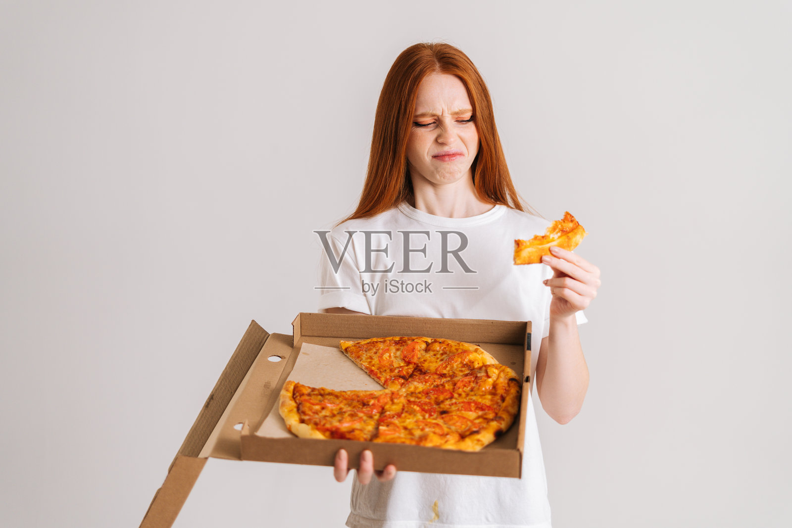 工作室肖像，沮丧的年轻女子吃坏质量披萨拿着盒子在白色孤立的背景厌恶地看着食物。照片摄影图片