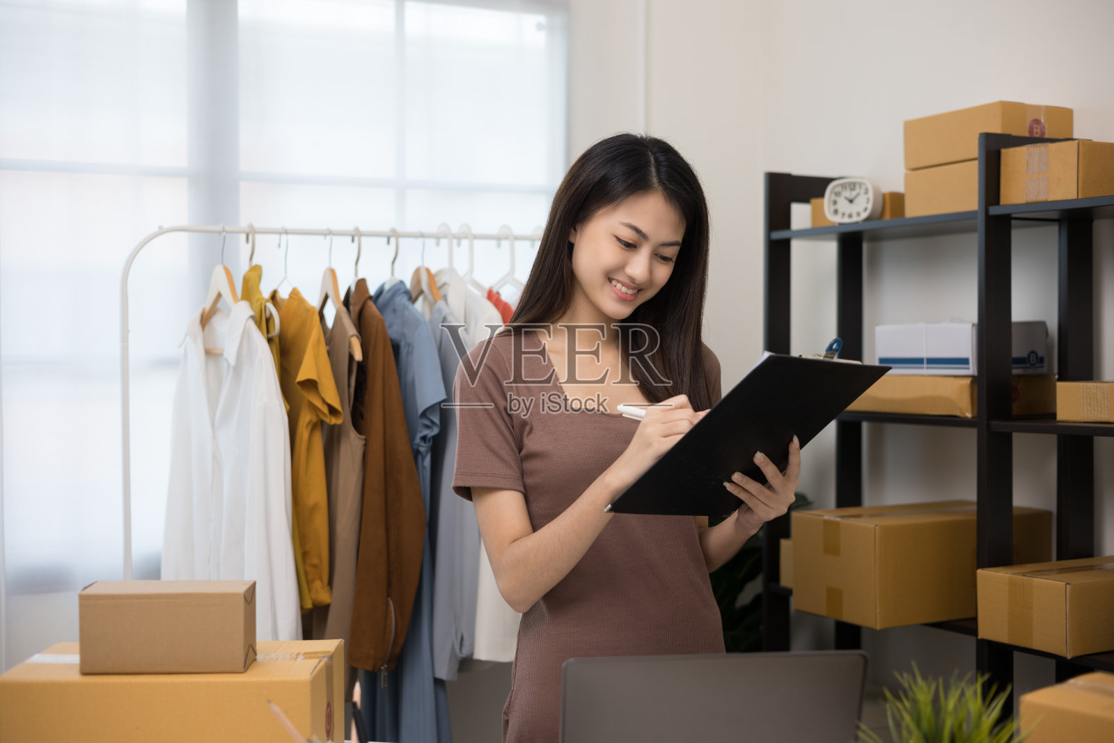 年轻美丽的亚洲女人在时装店老板持有剪贴板检查库存在轨道上的衣服。在服装店工作的亚洲女企业家。照片摄影图片