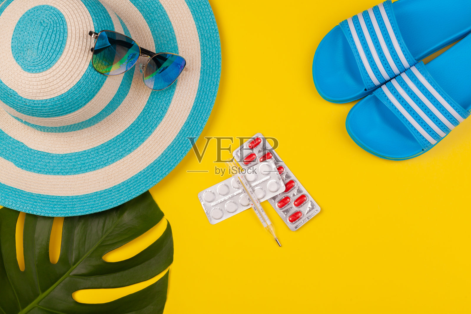 俯视图上的温度计，白色和红色药丸在水泡包装，蓝色帽子，蓝色人字拖，太阳镜，怪物叶在黄色背景。海滩度假、海上旅游、温暖阳光夏日的概念。适应环境，旅行期间的疾病，健康保险照片摄影图片