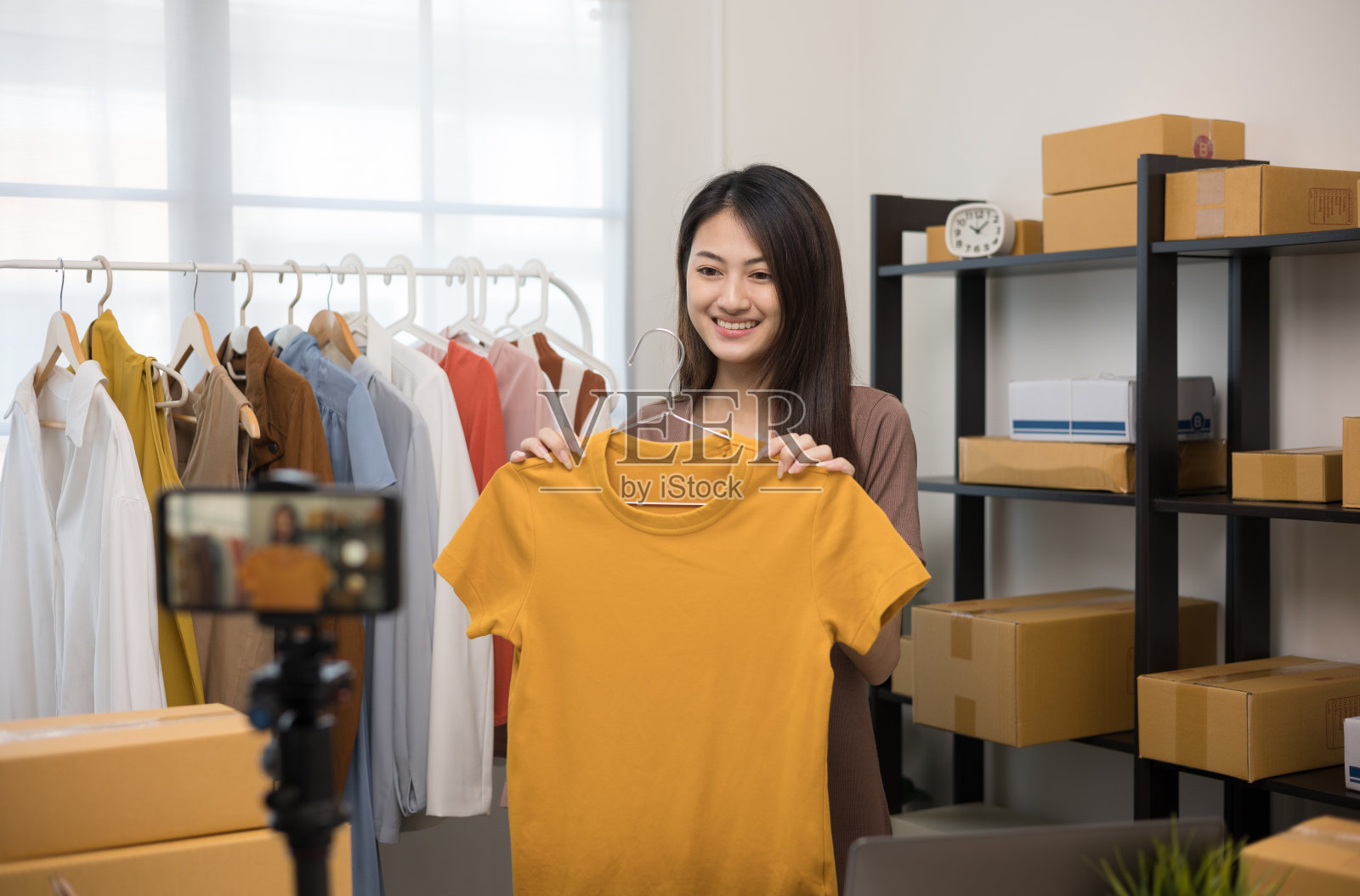 年轻的亚洲女性企业家在时装店使用智能手机直播销售时装服装。在社交媒体上出售的衣服，博主记录视频评论。网上购物的概念。照片摄影图片