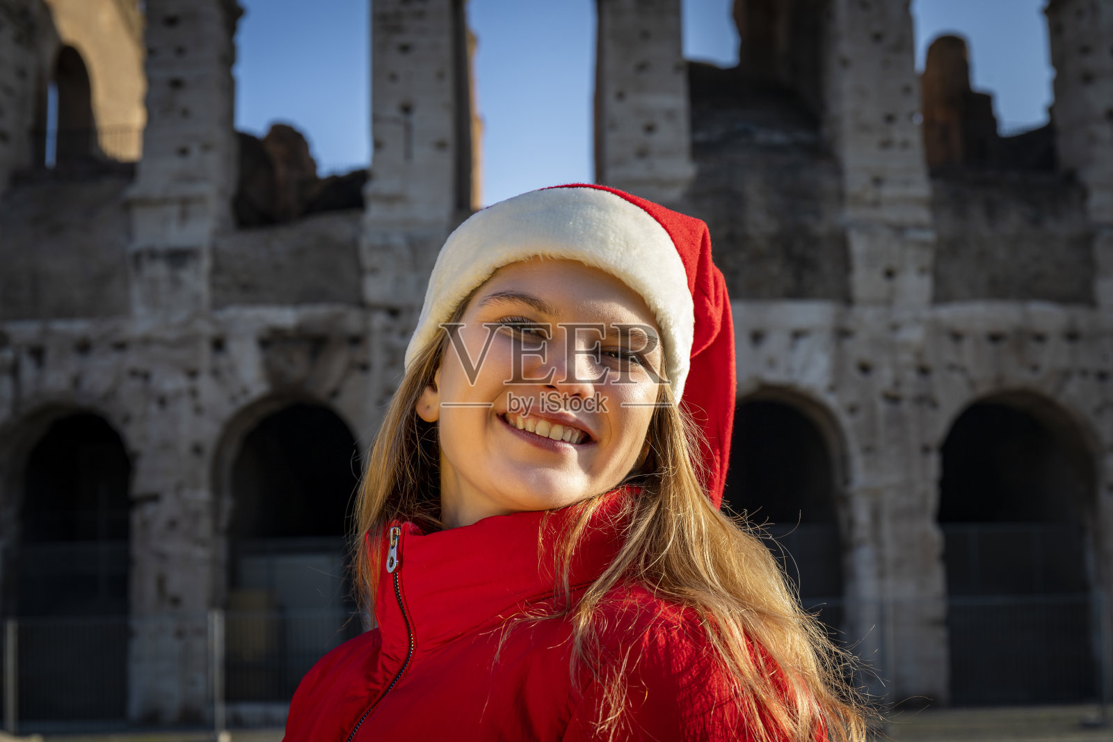 圣诞节是罗马假日。美丽的金发女郎在罗马圆形大剧场前摆姿势，戴着圣诞老人的帽子。照片摄影图片