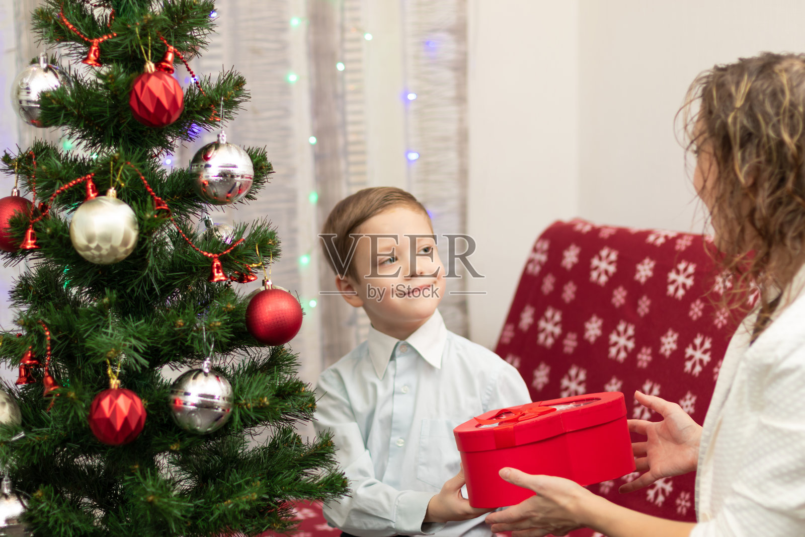 在家里的圣诞树背景下，一个穿着漂亮衬衫的7岁男孩给他的妈妈一个装在盒子里的新年礼物。有选择性的重点照片摄影图片