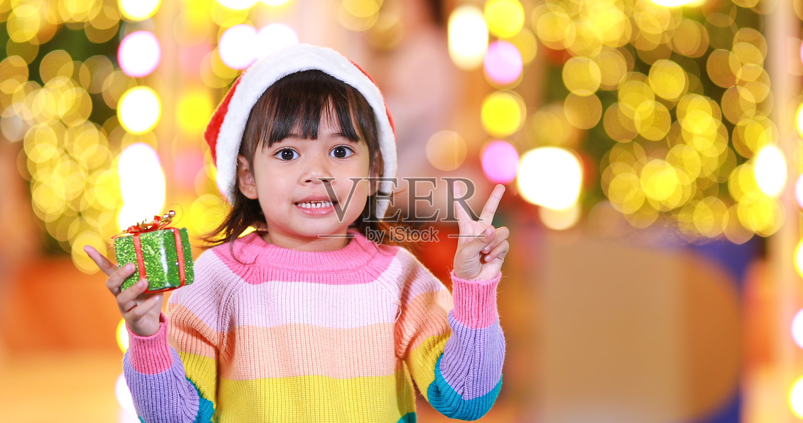 新年的期待。可爱的小女孩戴着红色的圣诞帽，穿着漂亮的彩色衬衫，背景是彩灯。在这个重要的季节里，尽情欢乐吧!照片摄影图片