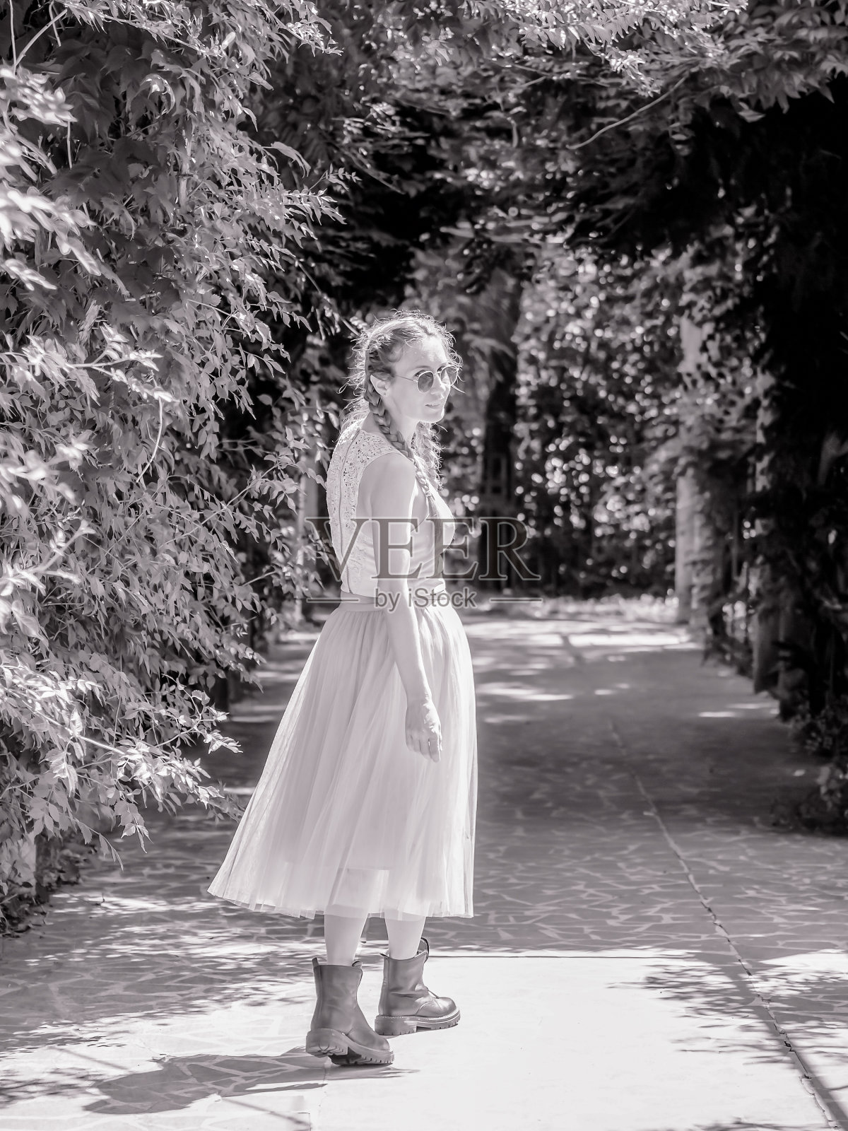 一个穿着薄纱裙子和粗糙靴子的女士走在公园的小巷里回头看。黑白照片照片摄影图片