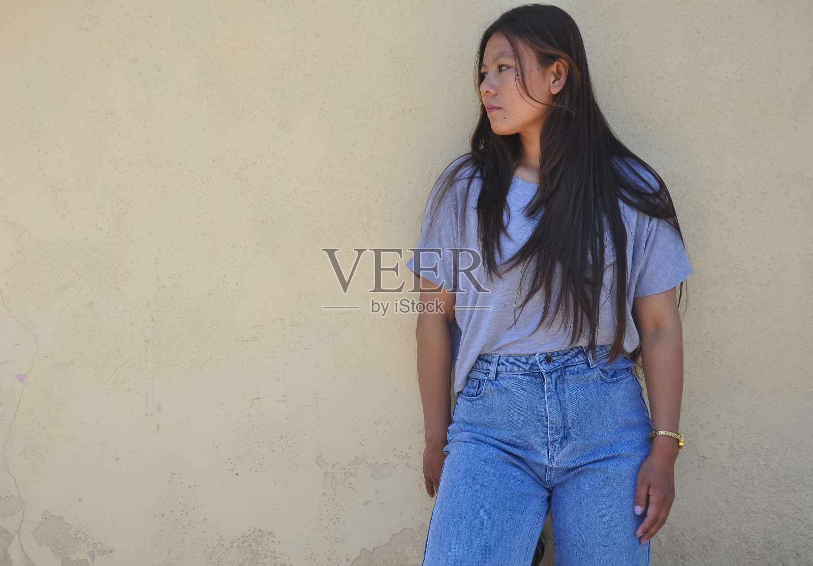 照片的南亚年轻女孩穿着灰色的t恤和蓝色的牛仔裤，站在黄色的墙与看她的右边与复制空间照片摄影图片