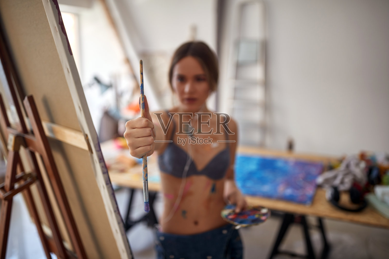 一位年轻性感迷人的女艺术家穿着胸罩站在画室的画架前，一边测量比例，一边画她的新画。艺术、绘画、工作室照片摄影图片