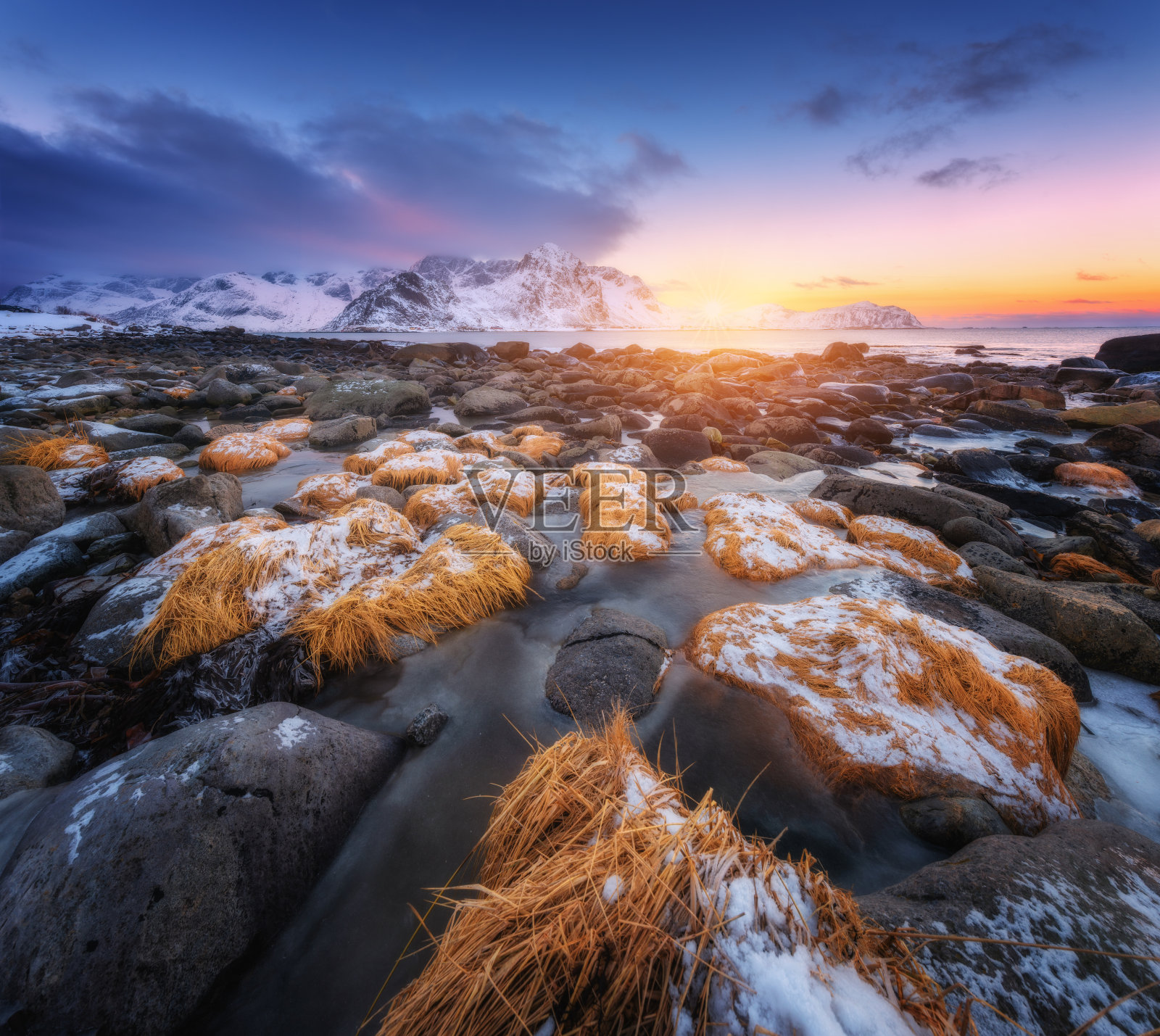 海滩上的石头和冰里的黄草，冬天日落时雪山、大海和彩色的天空。布满岩石的冰冻海岸。罗弗敦群岛,挪威。美丽的风景和海岸。自然照片摄影图片