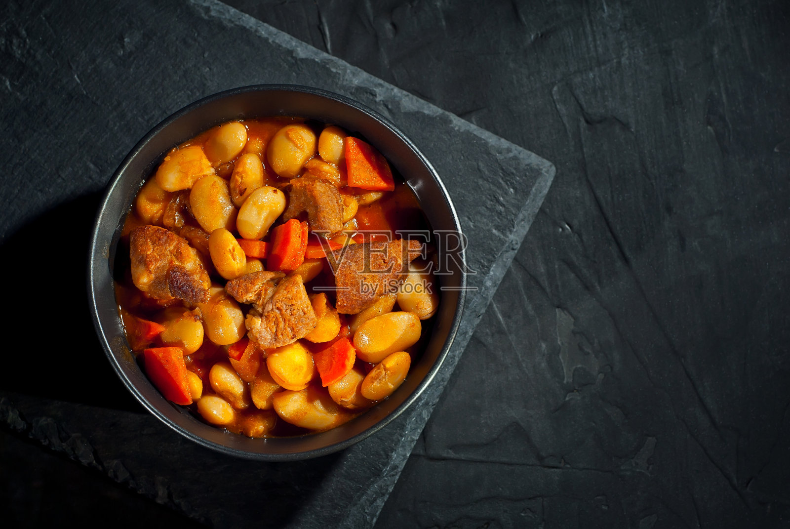 黑碗里的黄豆。煮豆子和肉放在石头桌上。豆子加番茄酱和猪肉。对比戏剧光线作为一种艺术效果。照片摄影图片