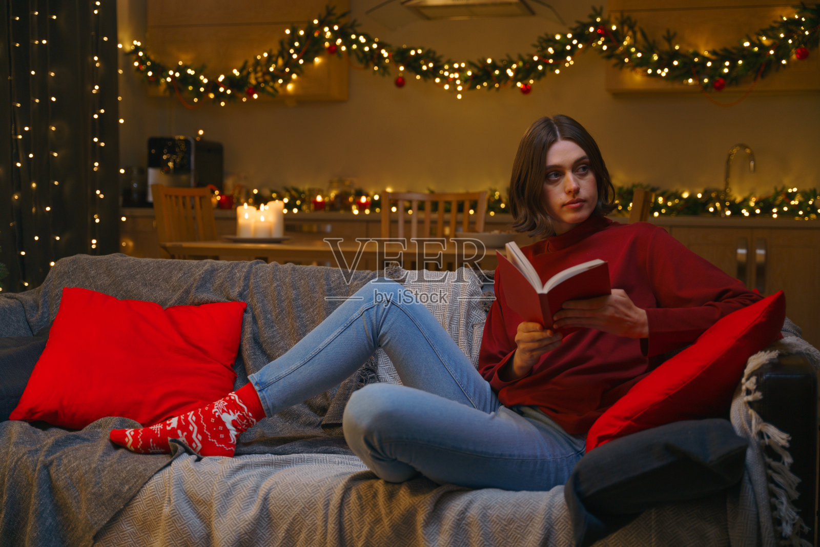 在圣诞节装饰的客厅里，一个年轻漂亮的女人正在阅读一本有趣的书，她坐在舒适的灰色沙发上，看着别处，想着她刚刚读到的东西照片摄影图片