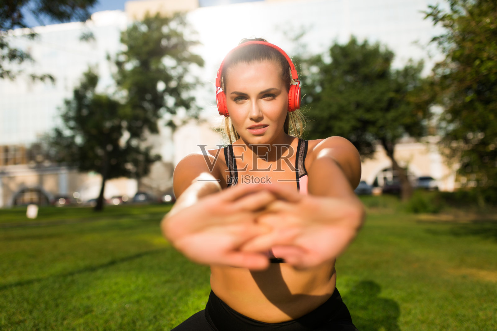 靠近年轻的加码女人在运动上衣与红色耳机伸展深思看在城市公园的相机照片摄影图片