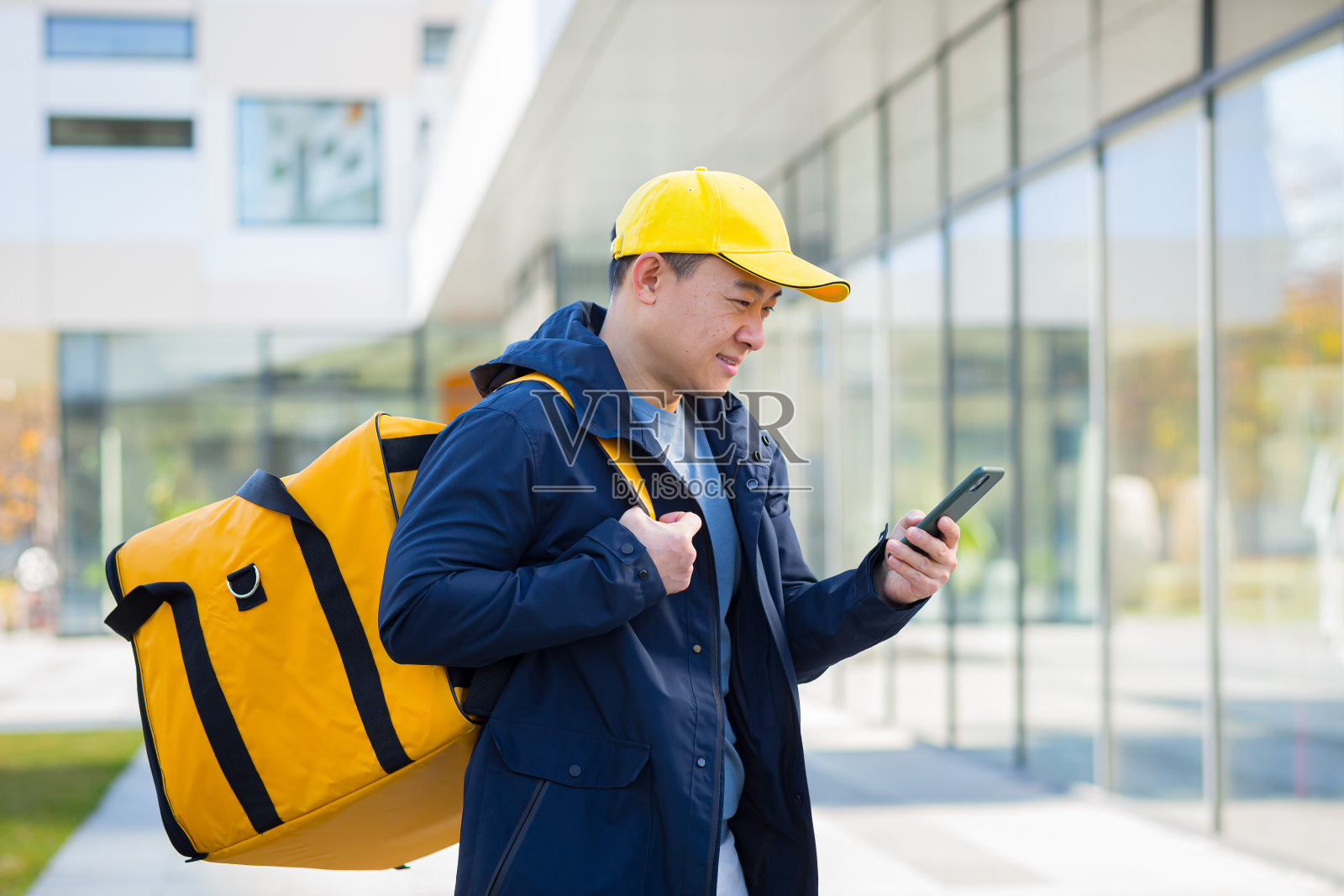 亚洲男性食品快递员使用手机使用手机应用程序在智能手机上，站在城市街道与隔热与热背包递送食品。客户地址。购买在线地图照片摄影图片