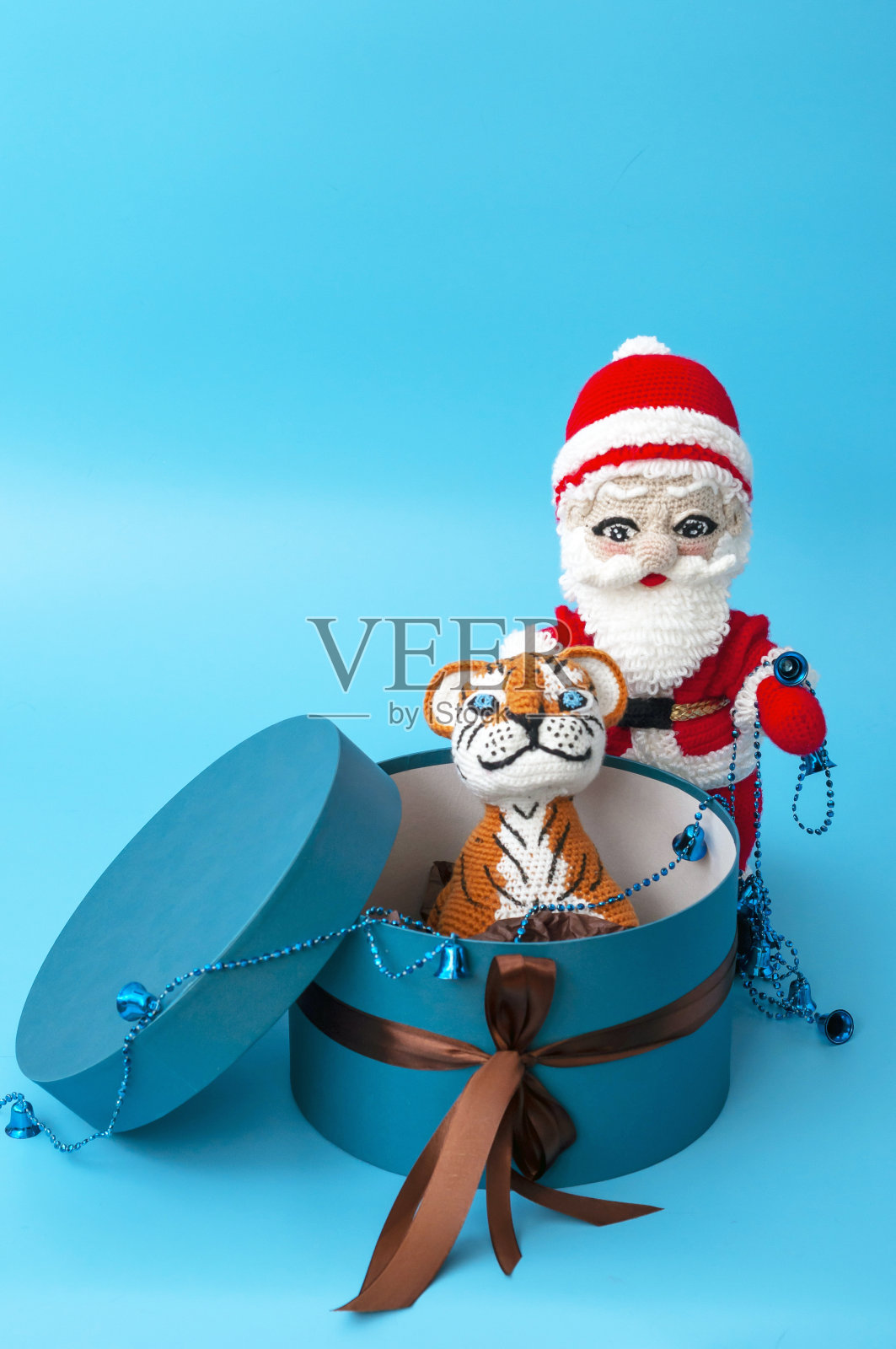 在蓝色背景上，一个编织的圣诞老人玩具站在一个蓝色礼品盒旁边，里面有一个编织的老虎玩具。垂直的快照。照片摄影图片