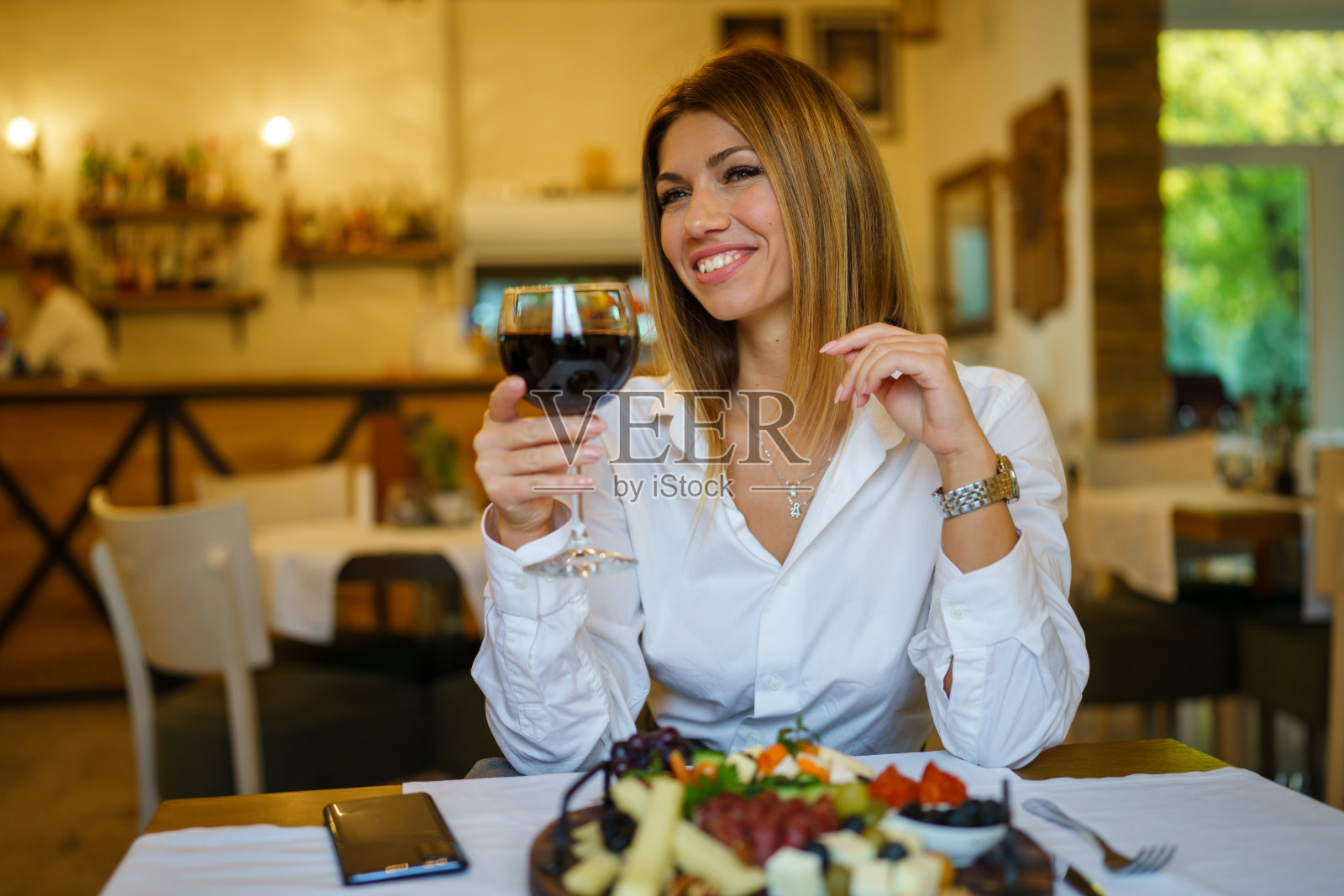 美丽的女人坐在餐厅里喝着红酒微笑着穿着白衬衫照片摄影图片