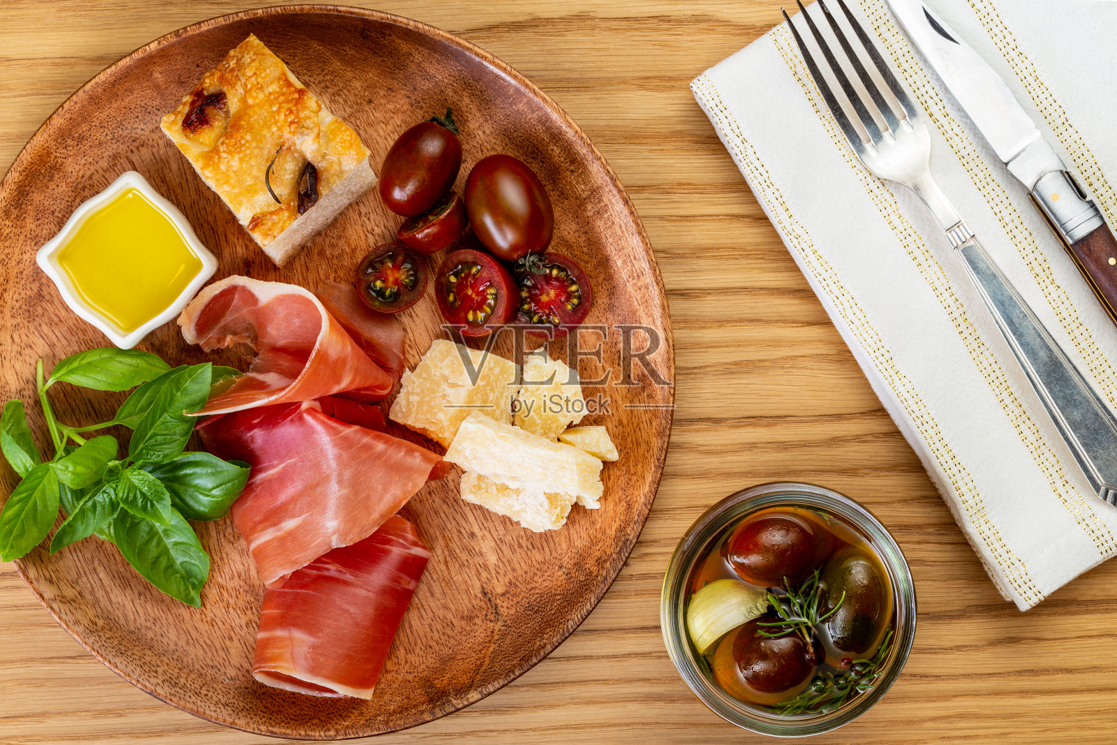 意大利火腿、帕玛森芝士、佛卡夏面包、番茄和罗勒枝放在木盘子里，旁边是白色餐巾，餐具以木头为背景。光意大利开胃小菜。平的。照片摄影图片