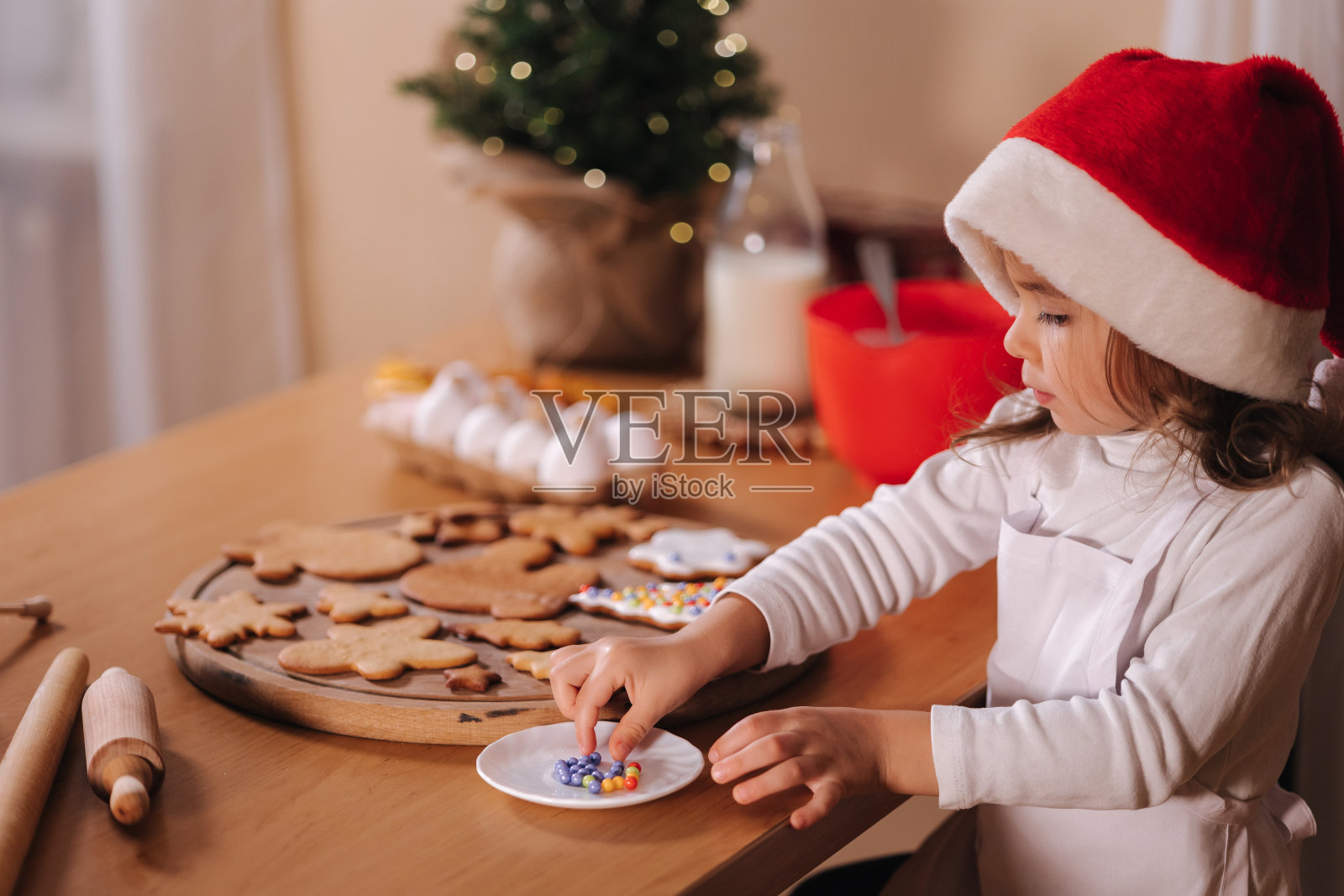 家庭烘焙，烹饪传统节日糖果。戴着红色圣诞帽的小女孩在木桌上准备做姜饼面团。新年庆祝传统。圣诞节的气氛照片摄影图片