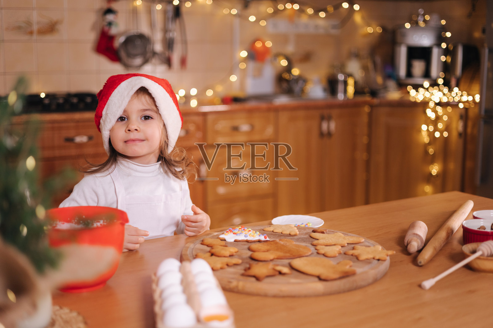 戴圣诞帽的小女孩用彩珠装饰姜饼。圣诞节和新年的传统概念。圣诞节的面包店。快乐霍利迪照片摄影图片
