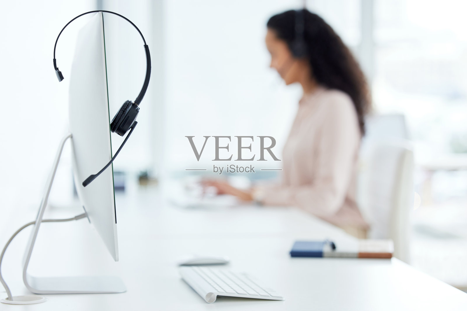 一个耳机放在电脑显示器上的特写镜头，一个呼叫中心的代理在后台工作照片摄影图片