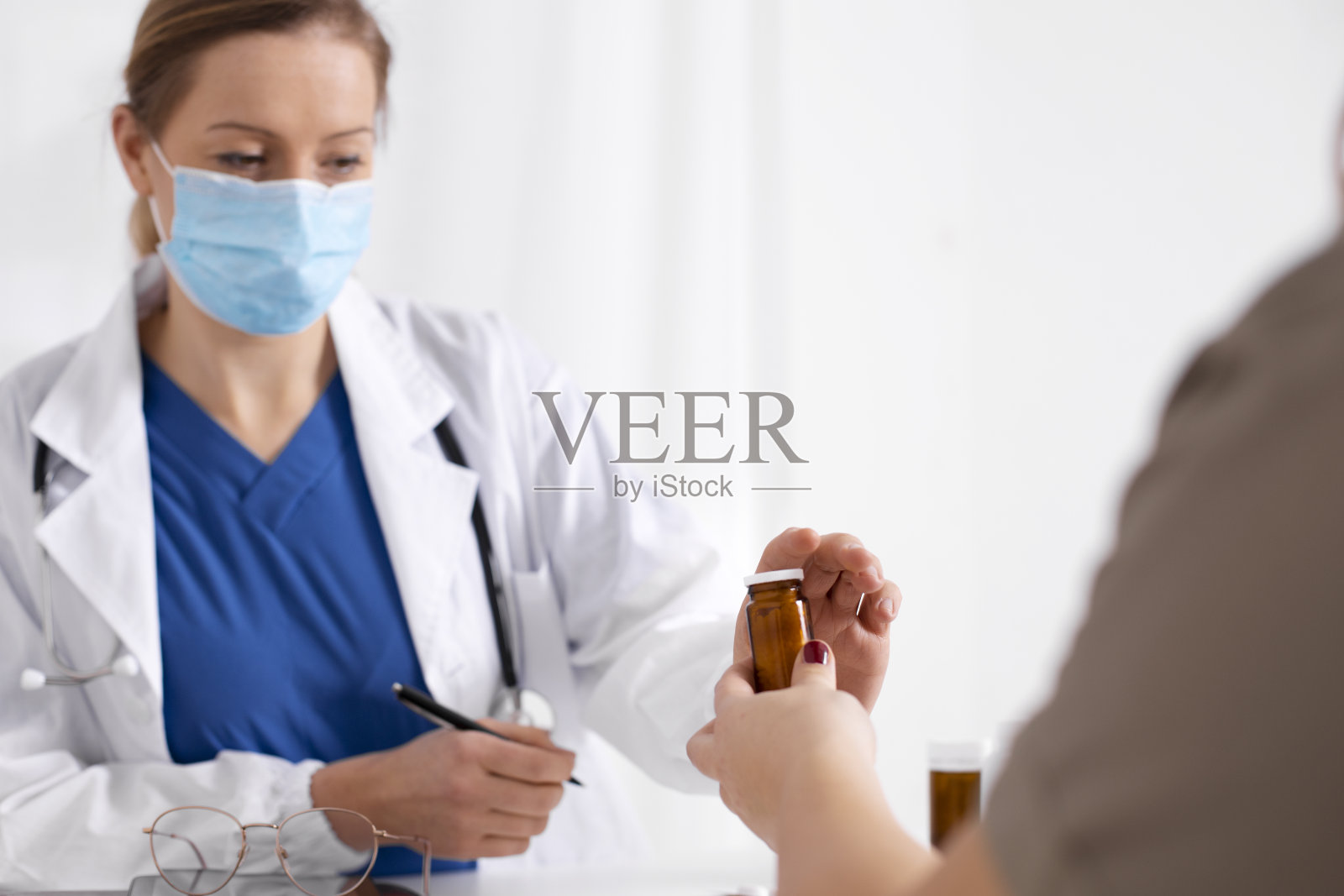 在冠状病毒和流感爆发期间，年轻的黑发女子在医生办公室咨询。女医生戴着口罩，给病人装药。病毒保护。Covid 19。照片摄影图片