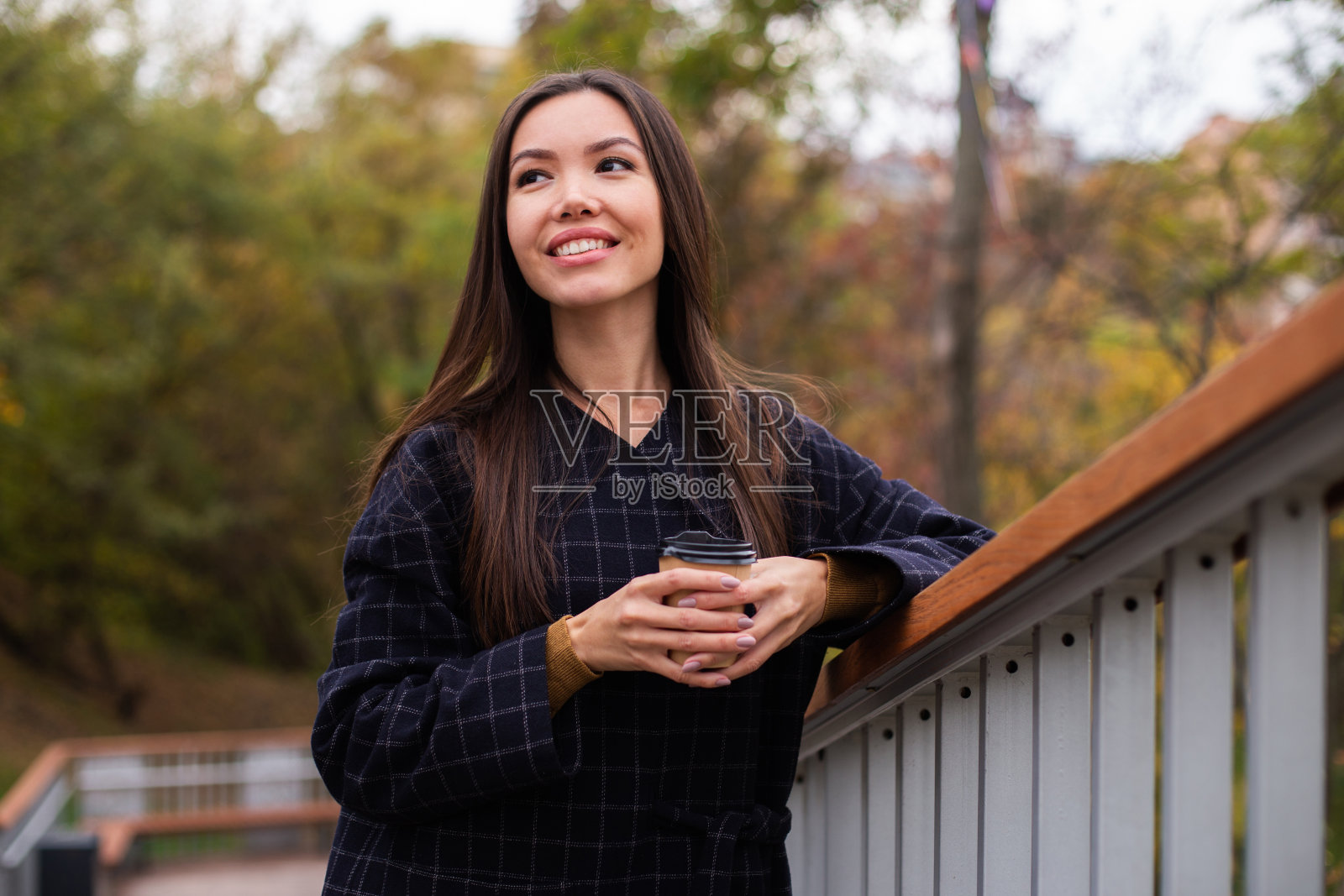 年轻漂亮的休闲女子穿着外套，喝着咖啡，在公园里开心地望着远方照片摄影图片