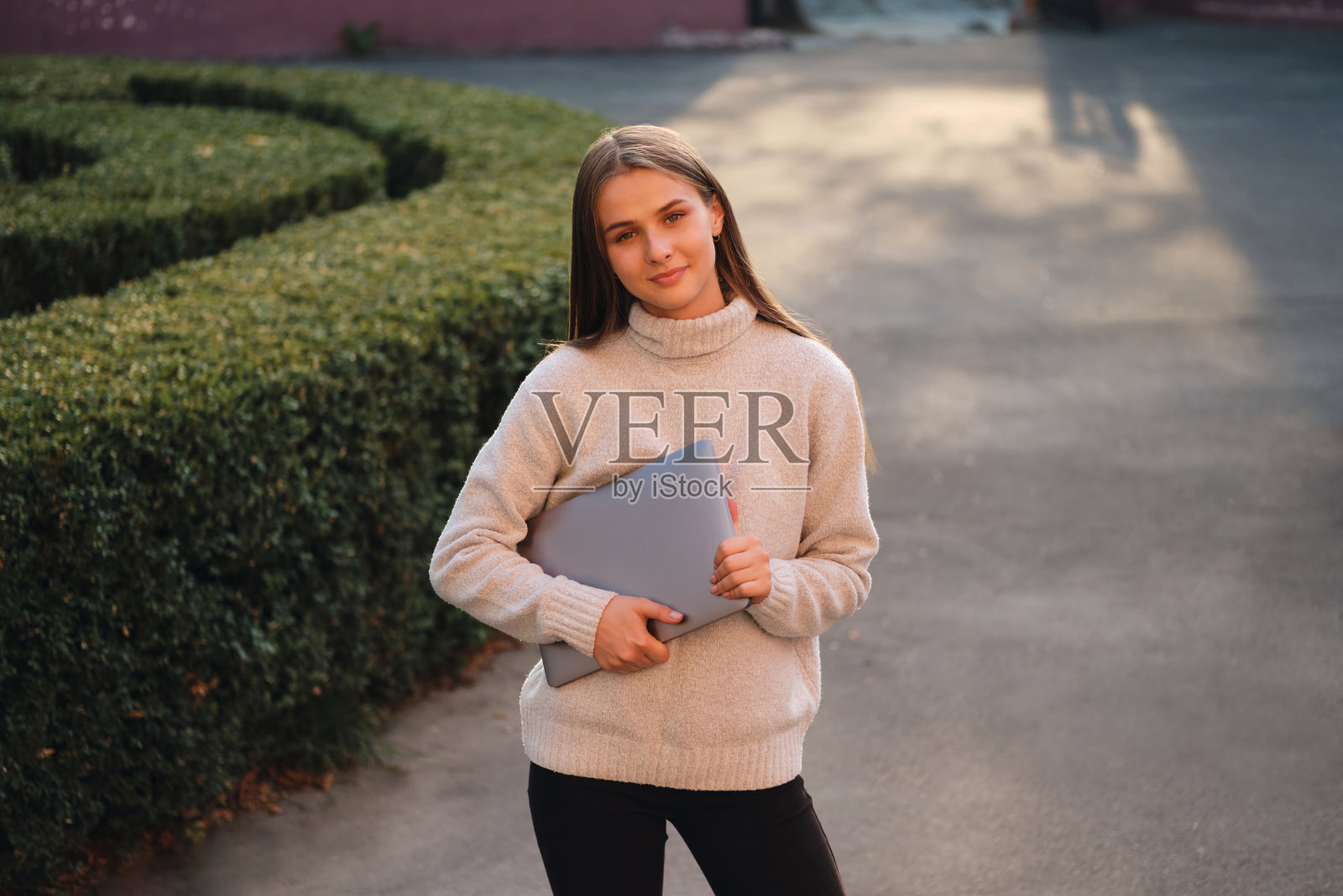 年轻漂亮的学生女孩穿着舒适的毛衣，带着笔记本电脑快乐地看着户外的相机照片摄影图片