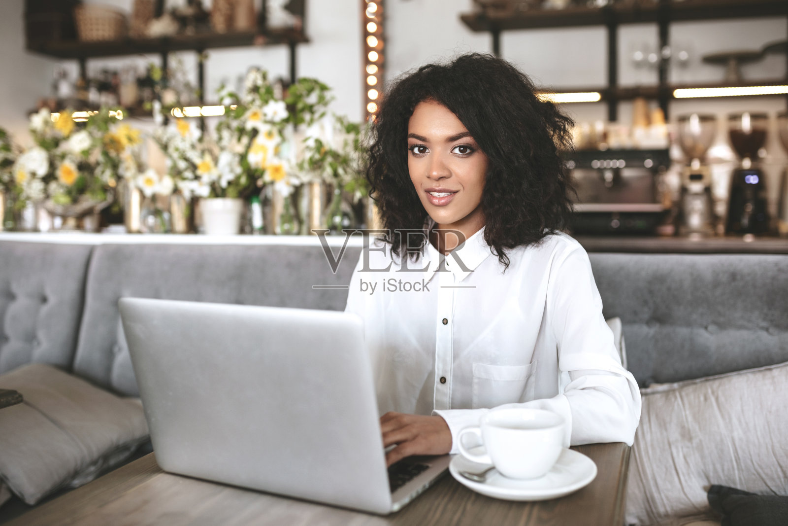年轻的非洲裔美国女孩坐在餐厅里，桌上放着笔记本电脑和咖啡。穿着白衬衫的漂亮女孩在咖啡厅用笔记本电脑工作照片摄影图片