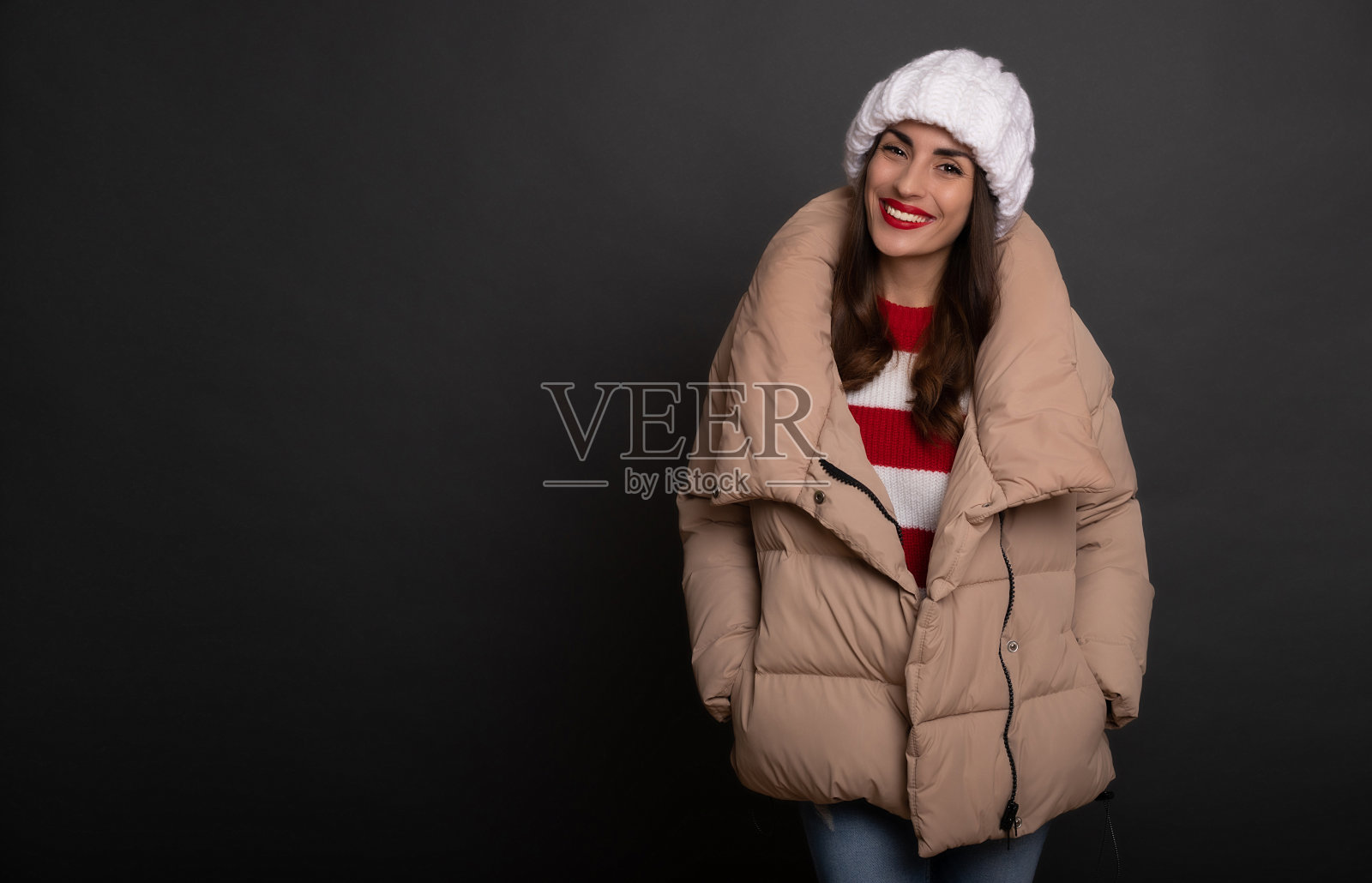 一个美丽的微笑的女人在冬天的帽子和温暖的时尚羽绒服的特写肖像，而她摆姿势，看着镜头孤立在灰色的背景照片摄影图片