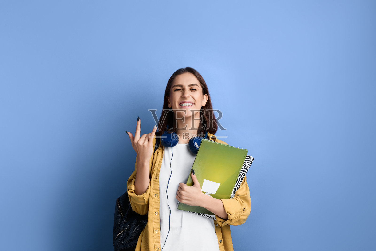 激动的女学生展示着摇滚的姿势，以优异的成绩通过了考试，为自己感到骄傲，拿着书，穿着黄色的衬衫，白色的t恤，黑色的包和脖子上的耳机。照片摄影图片