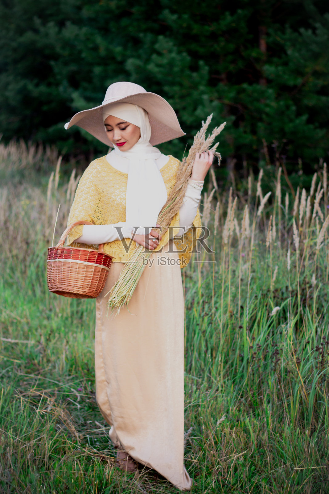 一位穆斯林妇女戴着帽子穿过田野，手里拿着小穗和柳条篮子。穿着米色头巾的穆斯林妇女。美丽,温柔,温柔。照片摄影图片