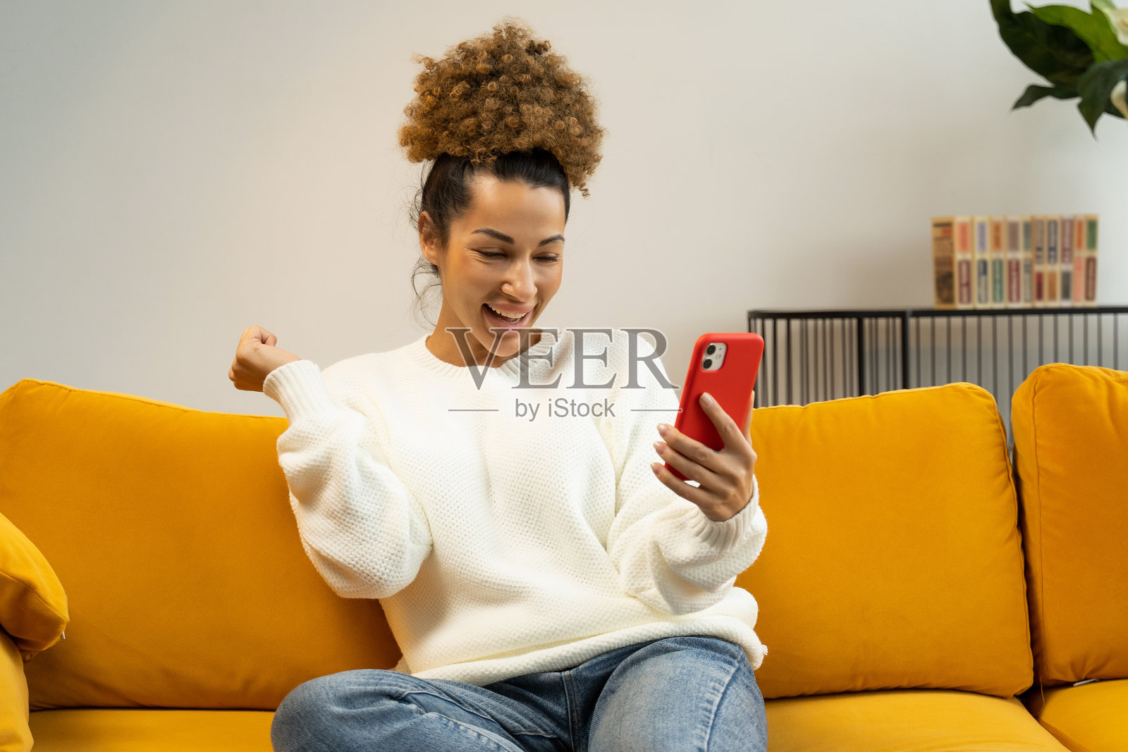 一位欣喜若狂的非洲裔美国妇女坐在家里的沙发上，用智能手机庆祝胜利照片摄影图片