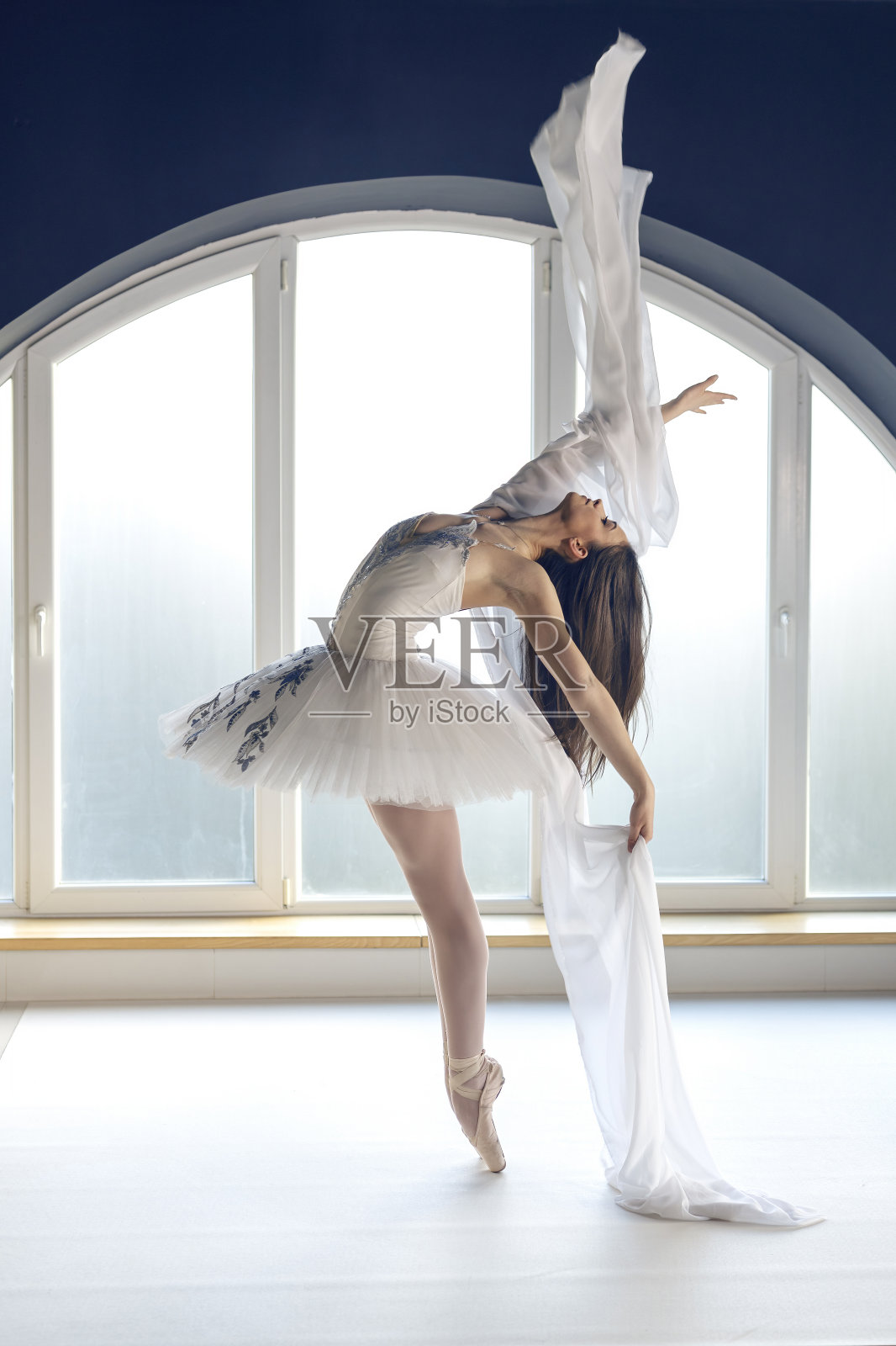年轻的芭蕾舞演员穿着白色的芭蕾舞服，在自然环境的日光下，在圆形大窗户背景的芭蕾舞室里练习芭蕾舞动作照片摄影图片