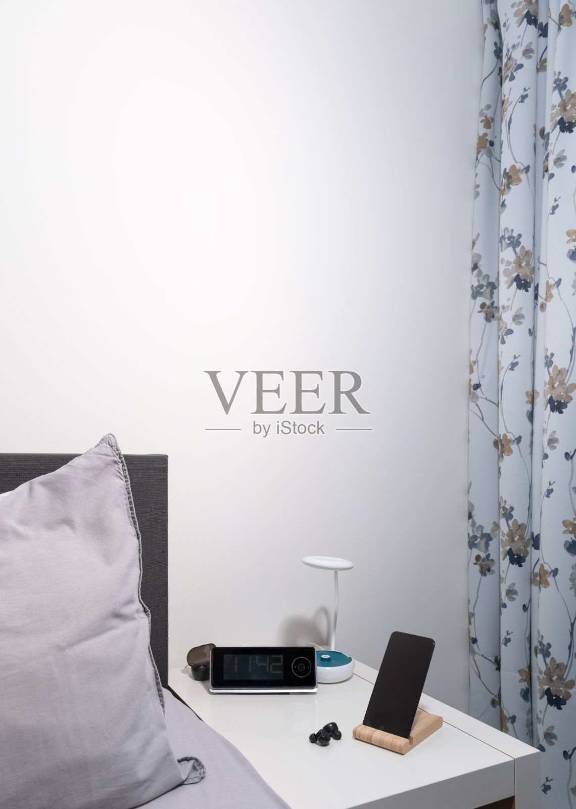 智能手机放在床头柜上，台灯、耳机、闹钟和床作为背景。垂直照片摄影图片