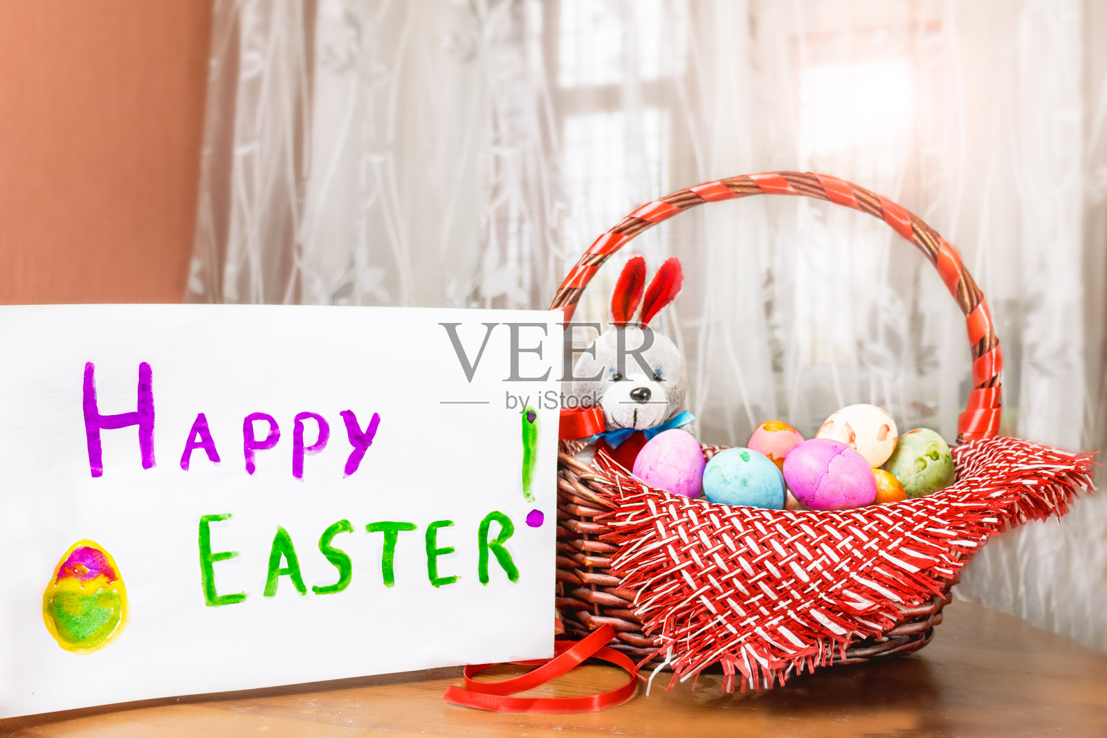 彩色的复活节彩蛋和一只玩具兔子躺在一个木篮子里，木桌靠窗，旁边是一张写着“复活节快乐”的白床单。选择性聚焦和白色背景。照片摄影图片