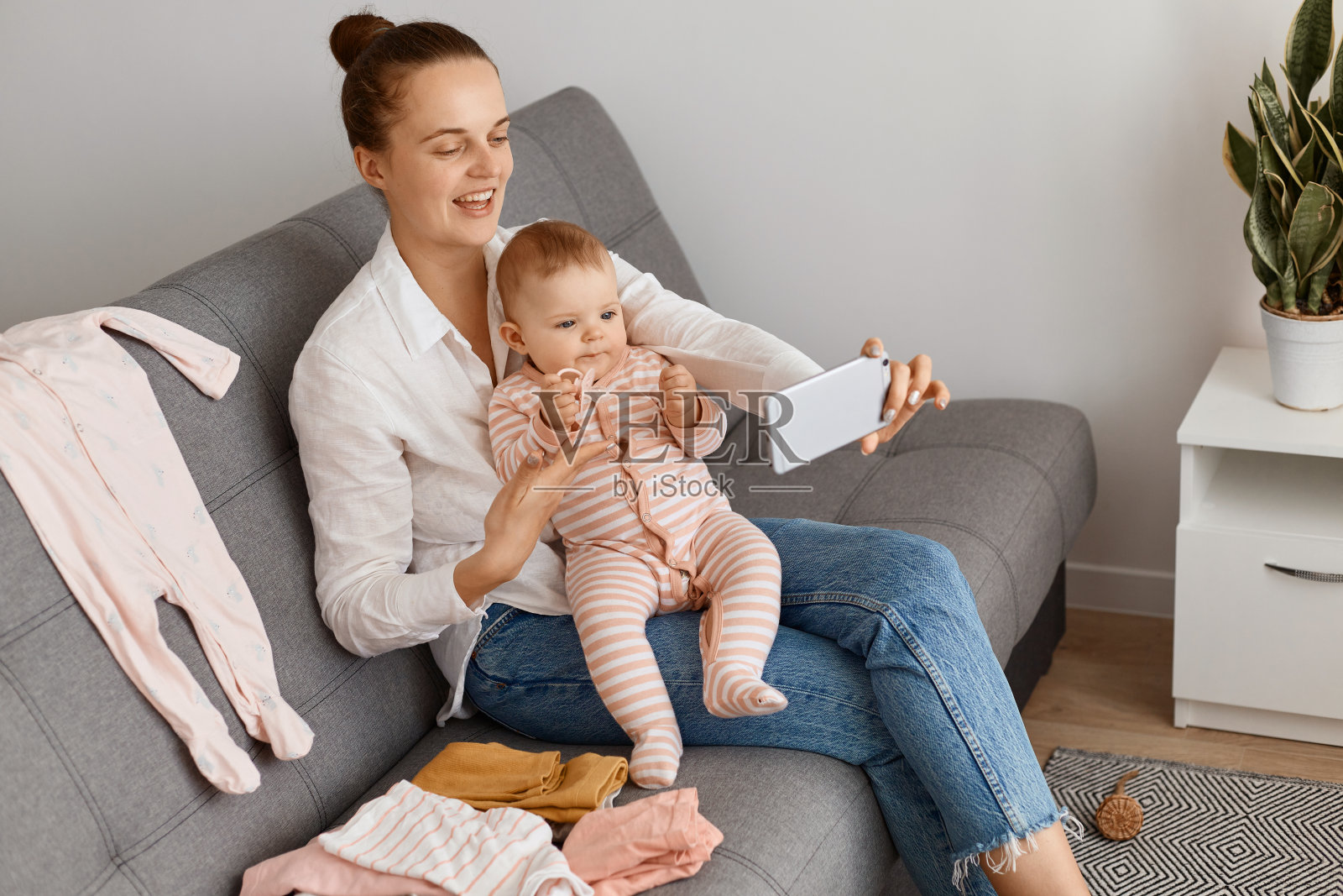 这是一个穿着白衬衫和牛仔裤微笑的女人，和她蹒跚学步的女儿坐在沙发上，和她的粉丝们聊天，表达积极的情绪。照片摄影图片