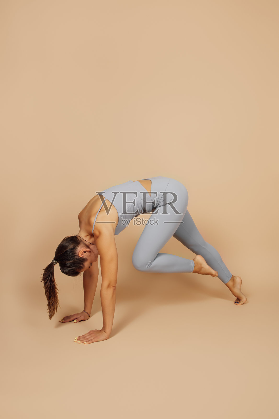 这位年轻女子正在做瑜伽。身着灰色运动紧身衣，头发扎成马尾辫的身材苗条的黑发女子，面朝下站着，双手撑地，膝盖抵胸照片摄影图片