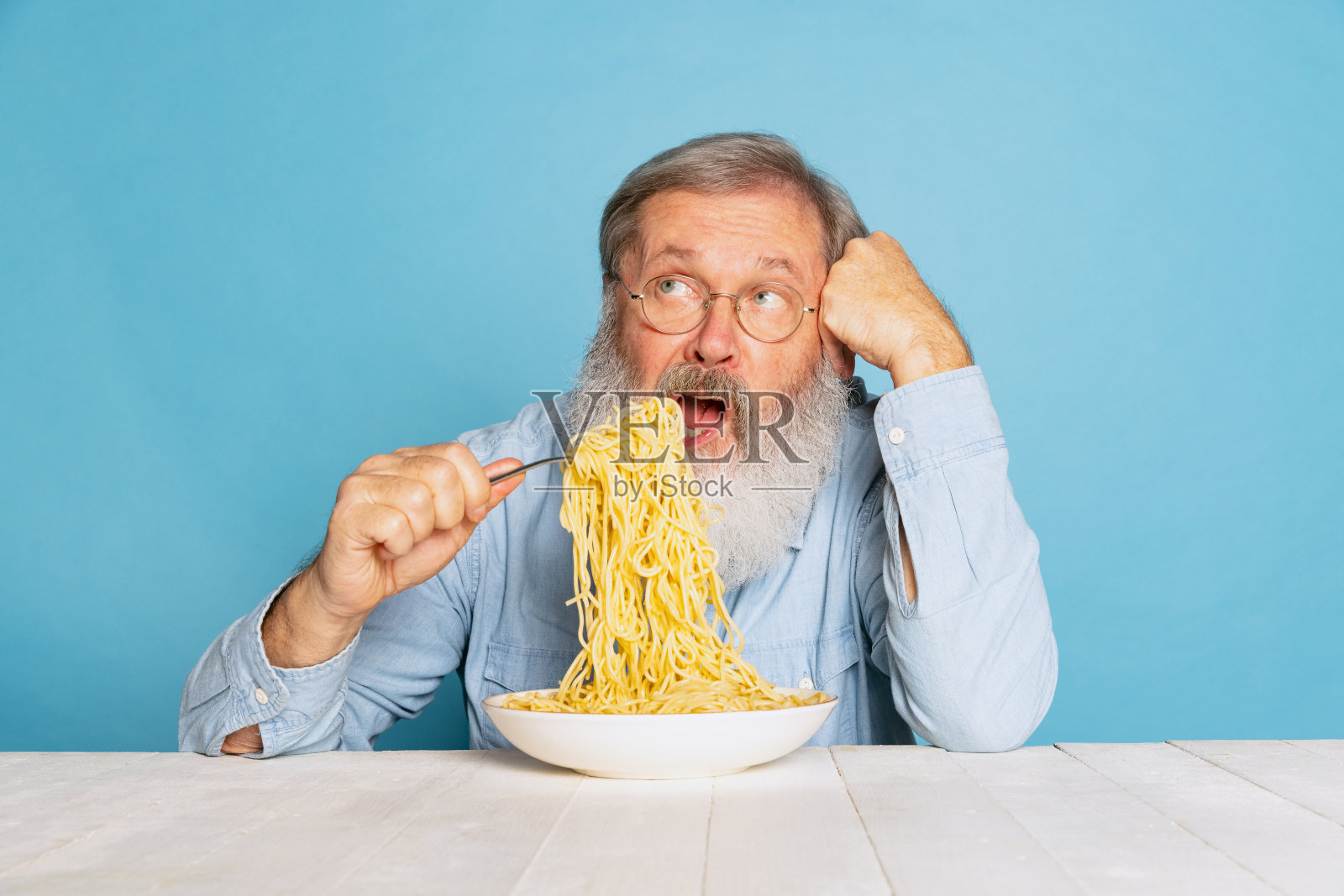 情绪激动的头发灰白的老男人吃着一大块面条，意大利面孤立在蓝色的工作室背景下。照片摄影图片