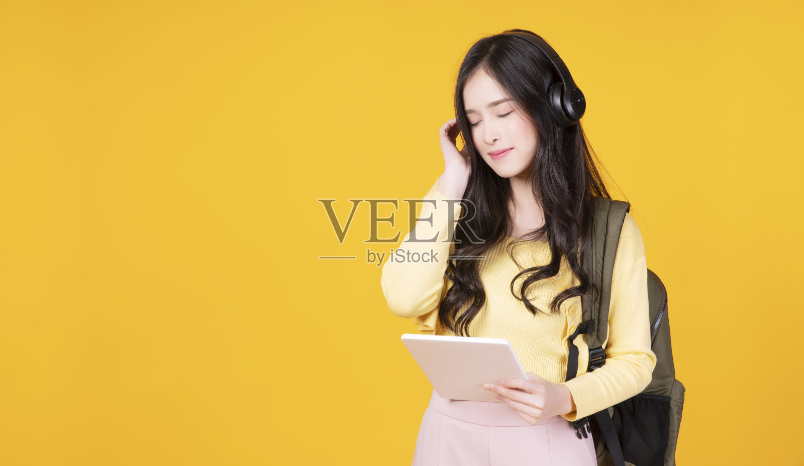 快乐的大学学生亚洲妇女持有平板电脑听课程在线与无线耳机在孤立的黄色背景。十几岁的亚洲女孩想一些放松积极的事情。照片摄影图片