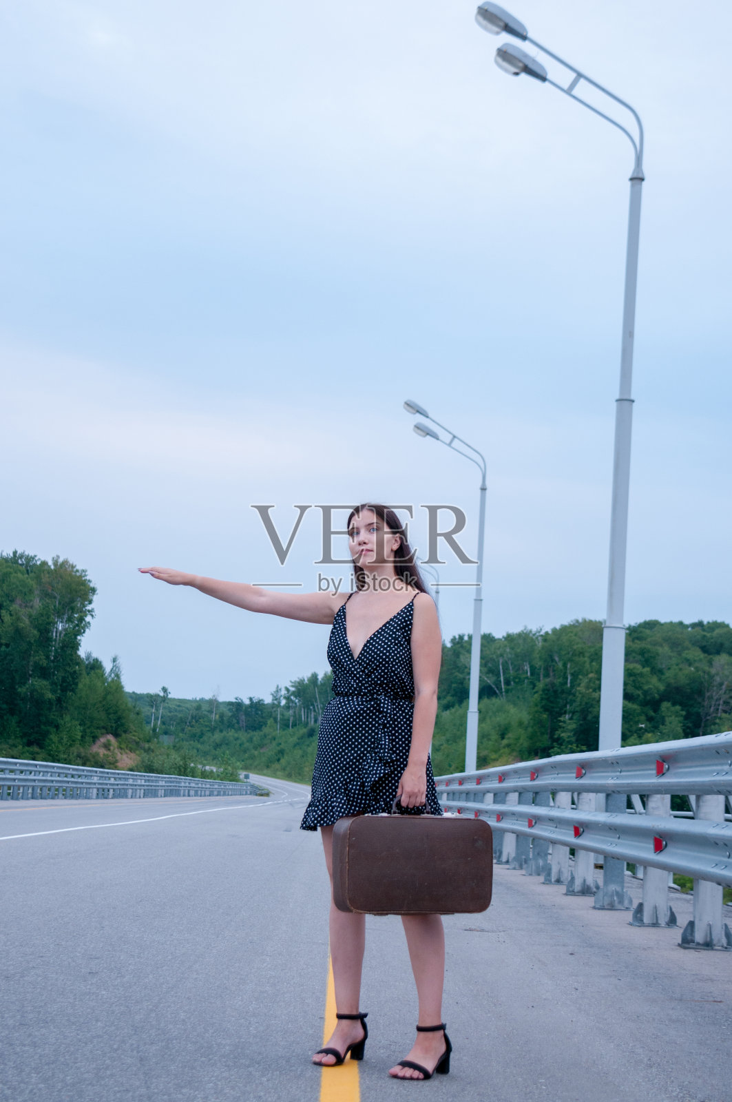 一个长着长发穿着黑色连衣裙的女孩带着一个手提箱在高速公路旁投票，搭便车的概念照片摄影图片