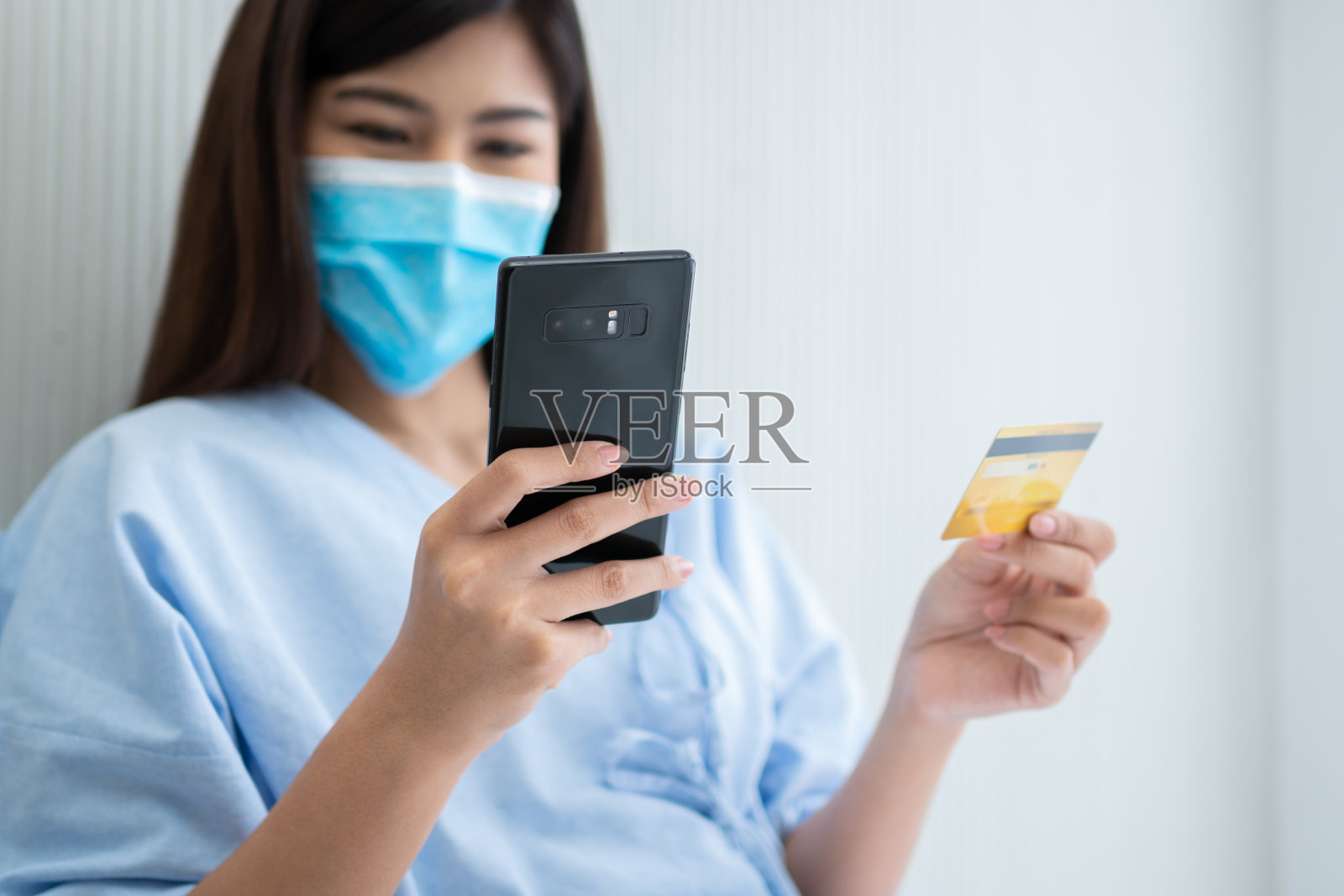 幸福的亚洲妇女戴着医用口罩，拿着假信用卡/保险卡和智能手机躺在医院的病床上，检查健康保险资格。银行保险，支付医疗照片摄影图片