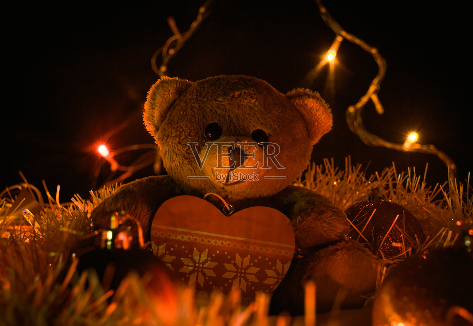 棕色泰迪熊坐在节日锡箔上的心形圣诞树玩具旁，被黄色花环照亮照片摄影图片