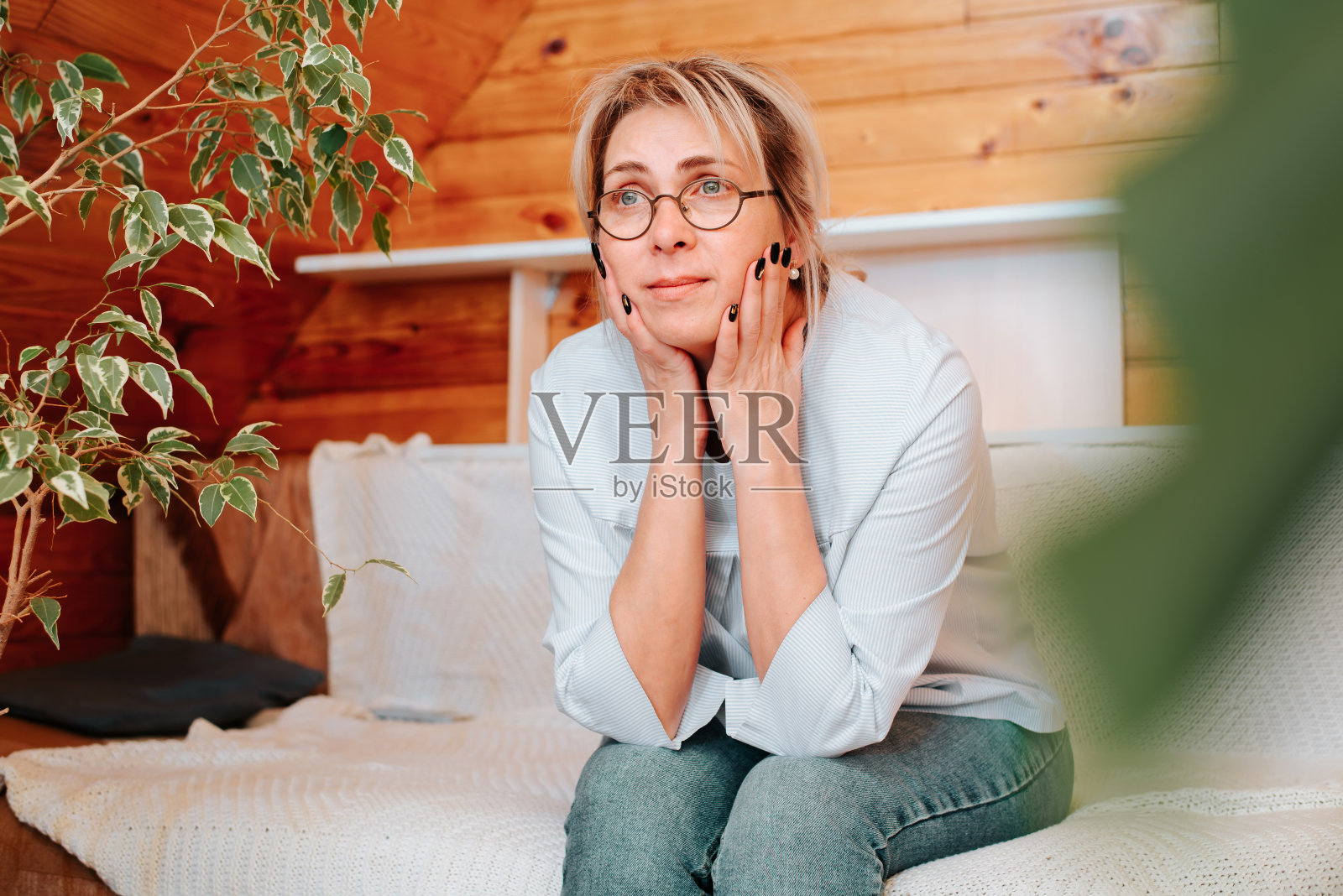 成年孤独悲伤的女人坐在沙发上，看着别处，在室内。中年白人妇女，戴着眼镜，穿着休闲的衣服独自在家。孤独、悲伤、压力的概念照片摄影图片