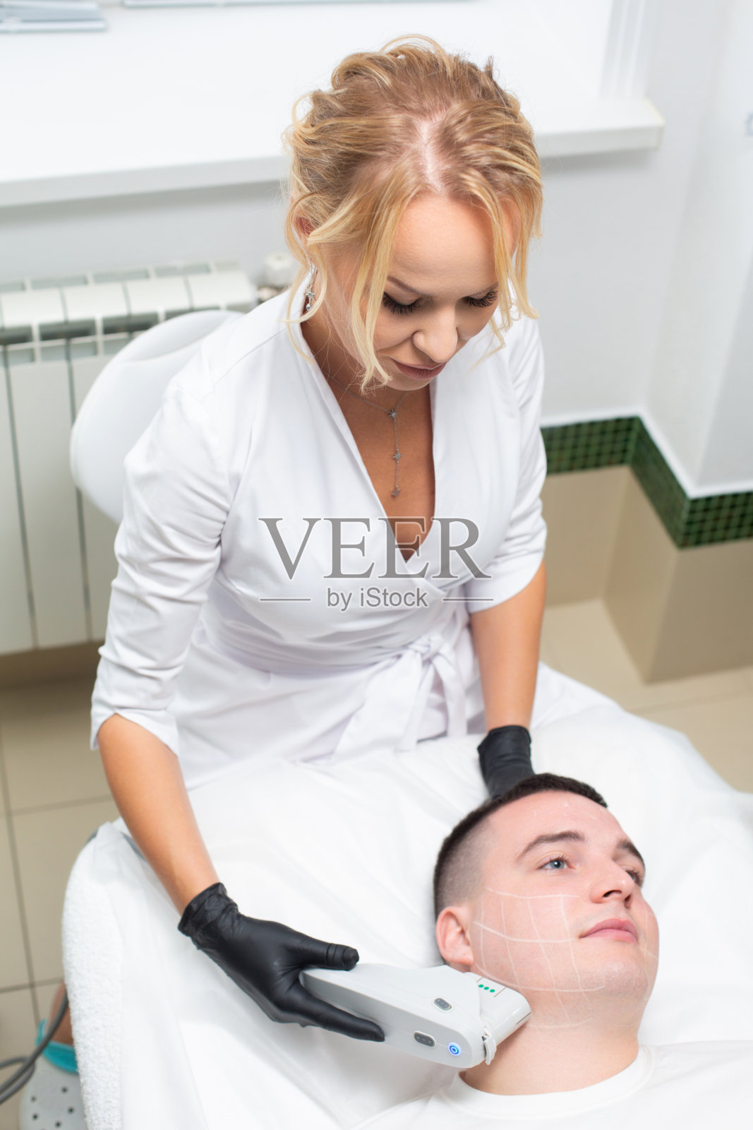 照片是一个年轻美丽的美容师进行激光面部毛发去除程序与现代设备的一个男人躺在沙发在美容办公室照片摄影图片