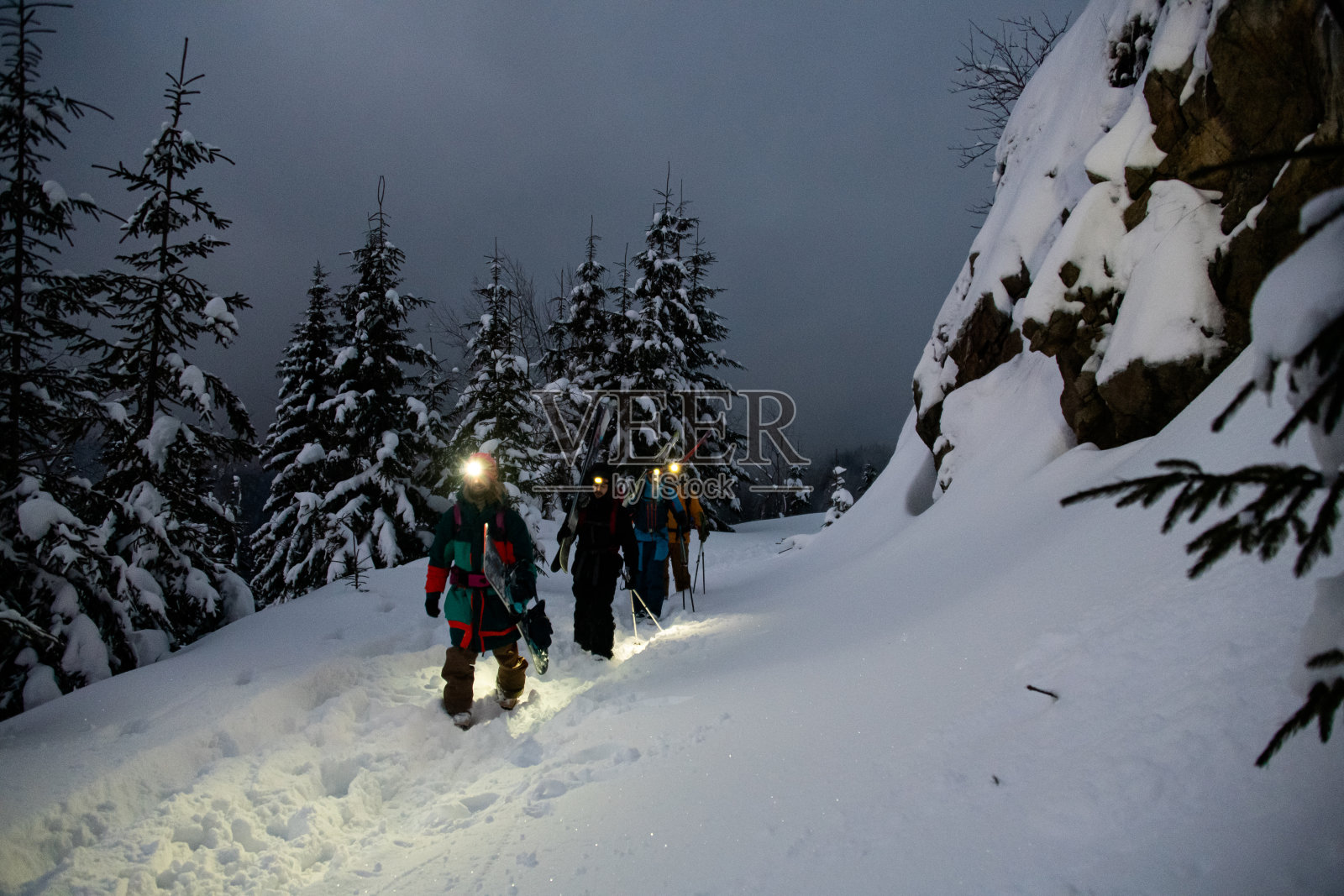 一群男性滑雪者走在雪山上的小道上，背景是白雪覆盖的冷杉树照片摄影图片