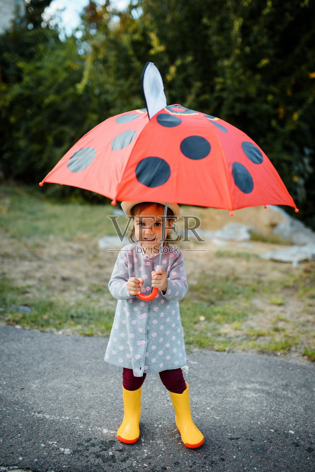 快乐的微笑的2岁小女孩穿着黄色雨靴，正在户外玩她的红色圆点伞。照片摄影图片