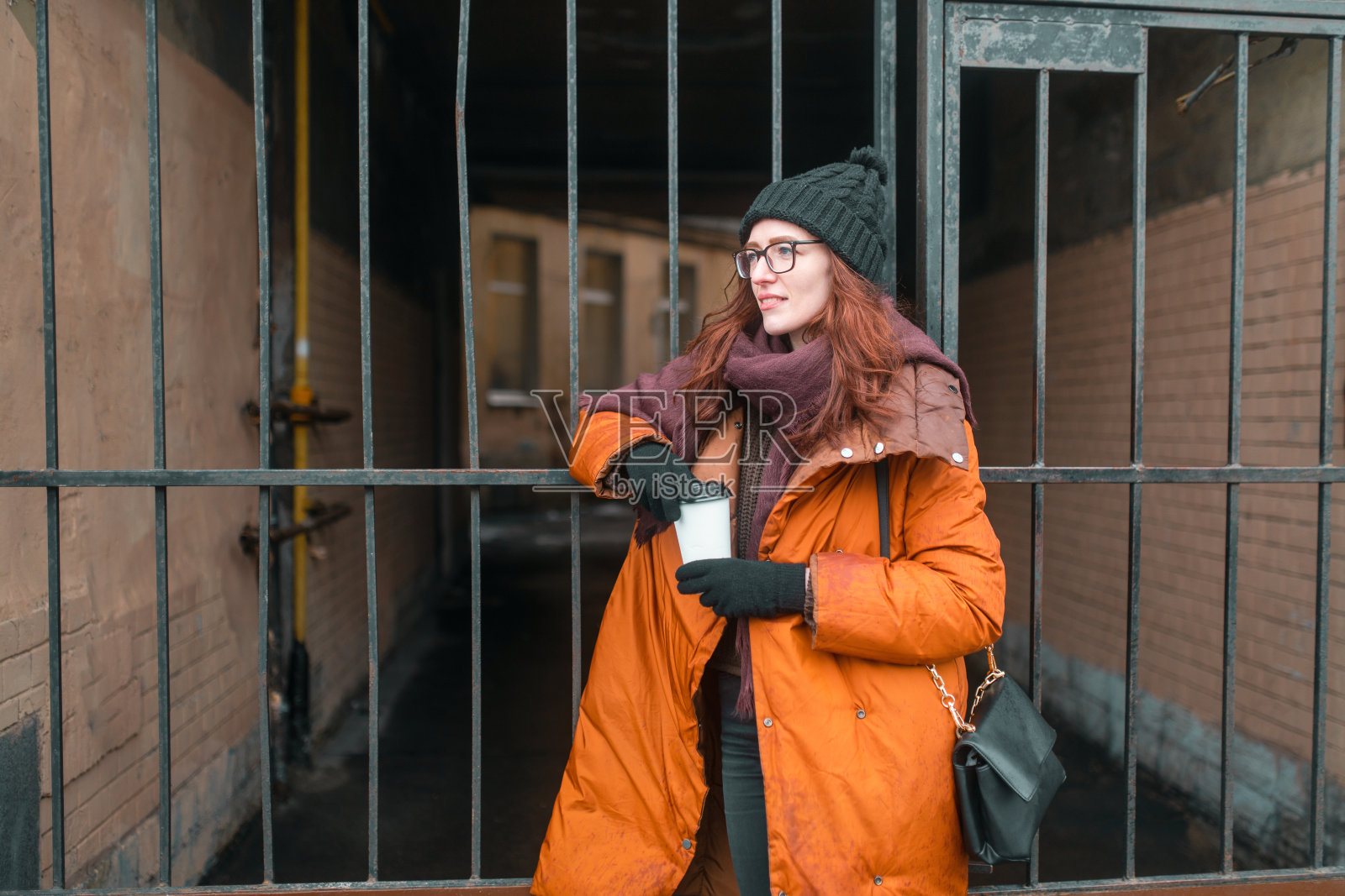 在寒冷的秋天，一个女人用热咖啡取暖。戴着眼镜。暖和的冬衣，帽子和围巾。照片摄影图片