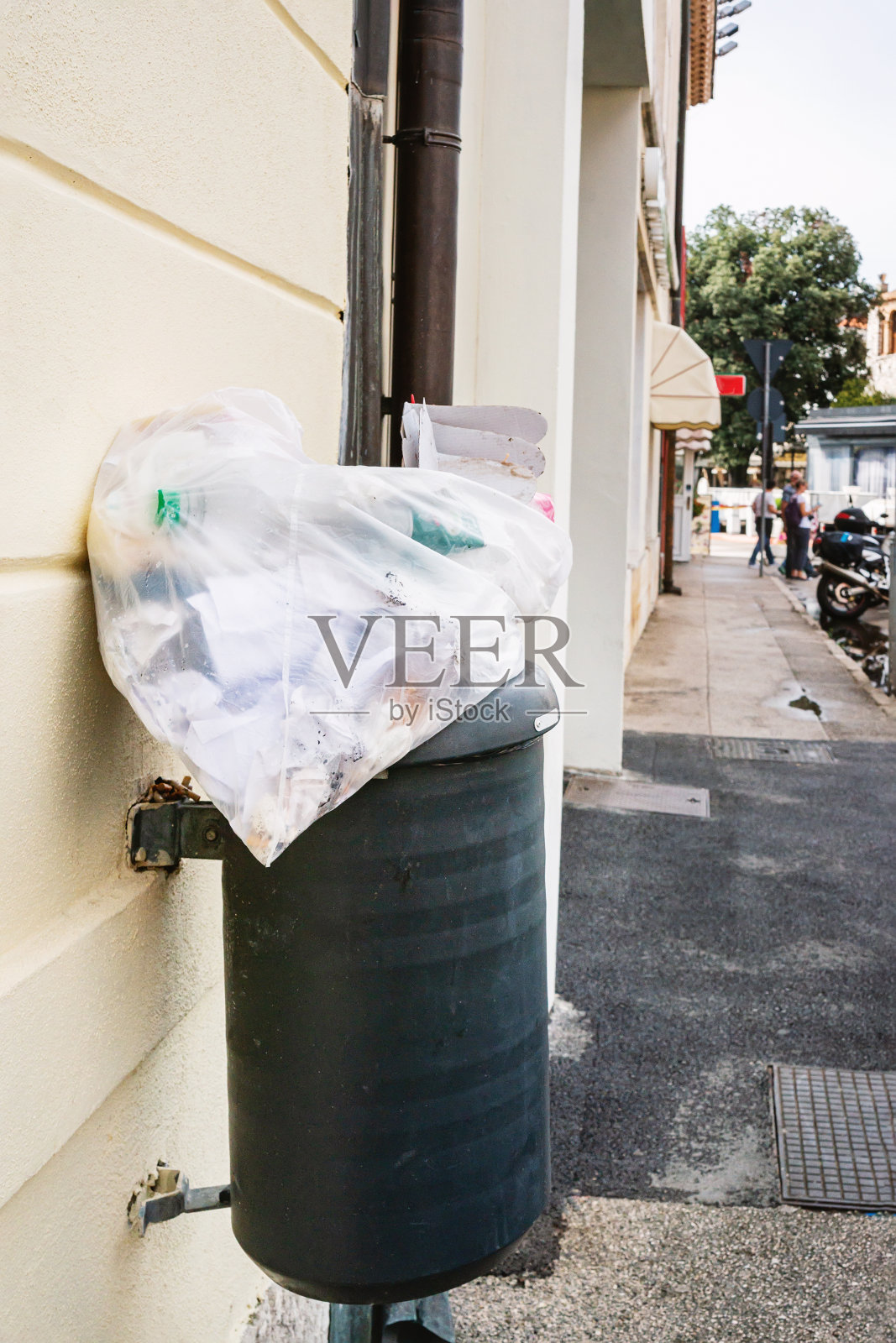 黑色的垃圾桶在街道的背景上放着一袋垃圾。一个满溢的垃圾桶靠在砖墙上。现代世界中塑料和垃圾的过剩照片摄影图片