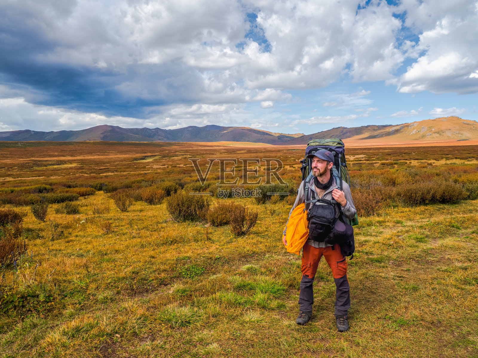 疲惫的游客背着大背包，身体两侧都是背包，在苔原山脉的背景下，黑色的戏剧性的云朵衬托下，他愉快地望着远方。照片摄影图片