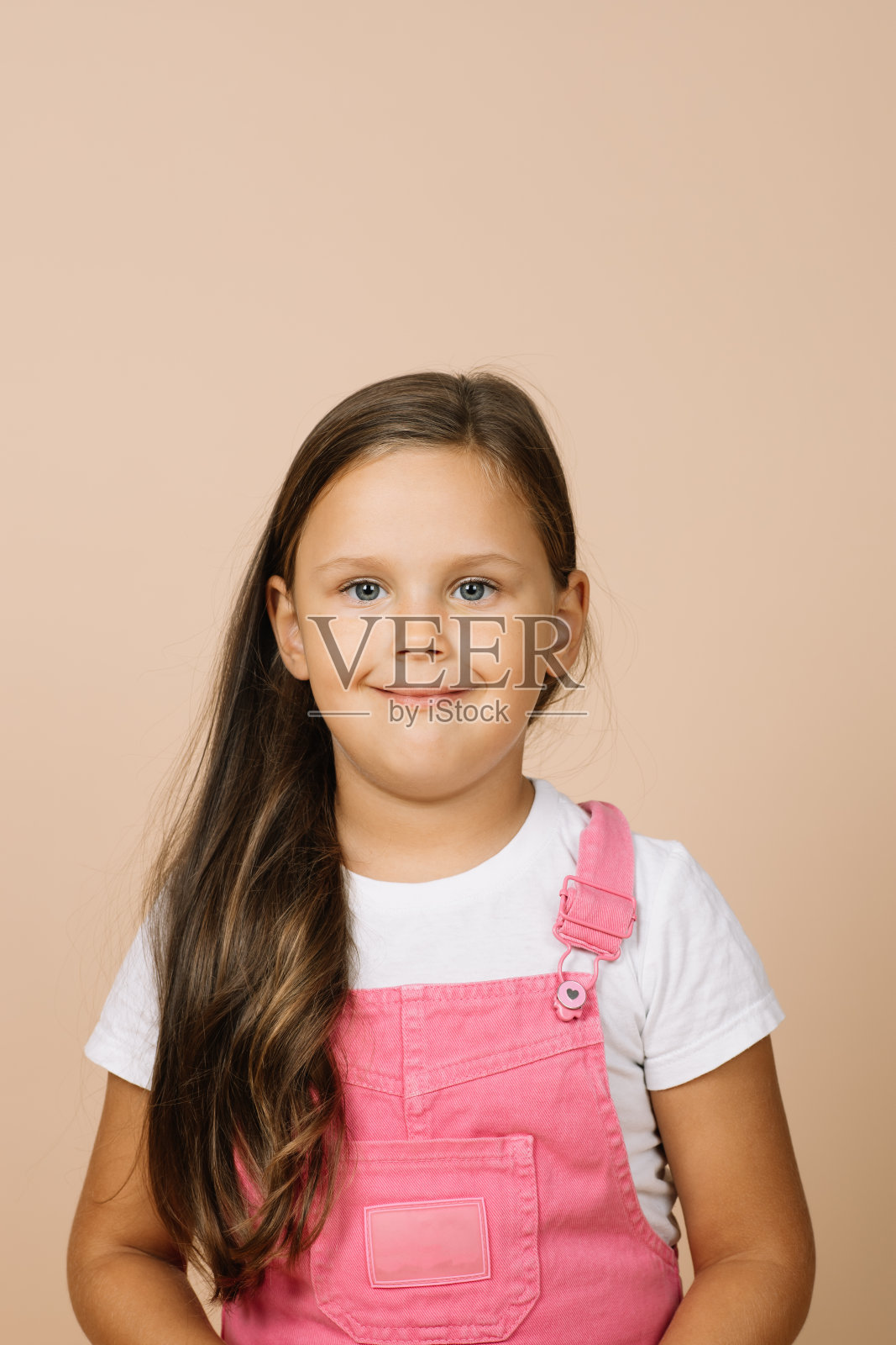 近脸孩子明亮的闪亮的眼睛和平静的快乐的宽微笑看着镜头穿着亮粉色的连体衣和白色的t恤在米色的背景照片摄影图片