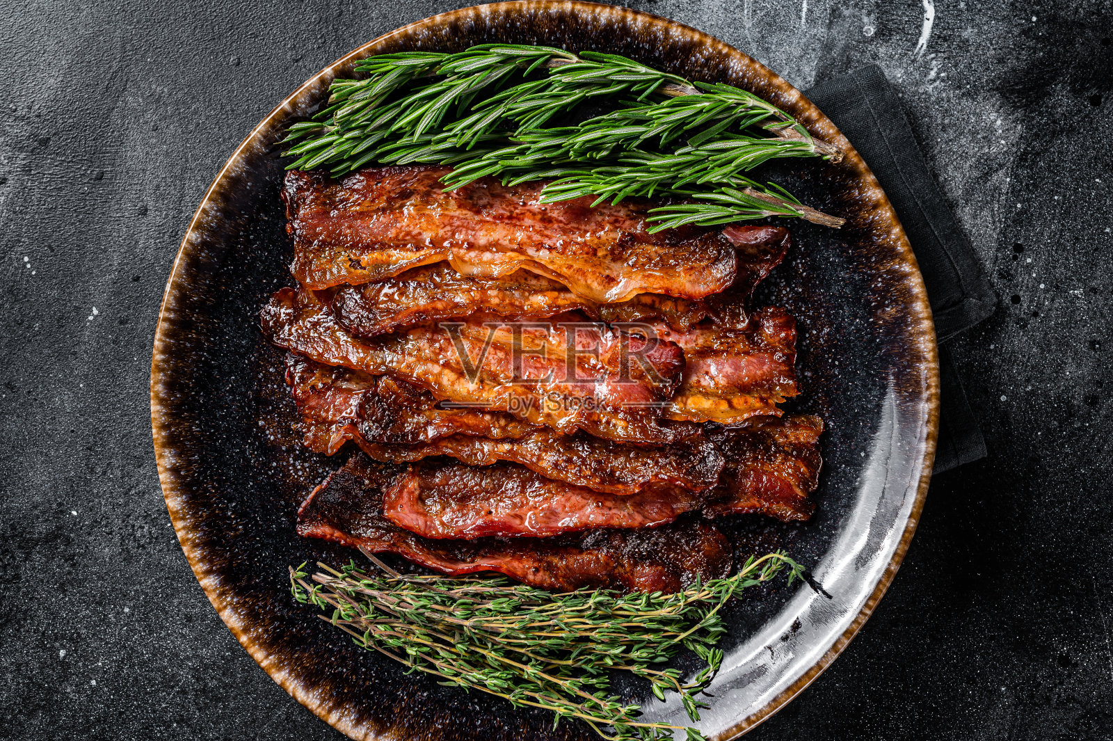 热炸脆培根片与香草在盘子里滋滋作响。黑色背景。俯视图照片摄影图片