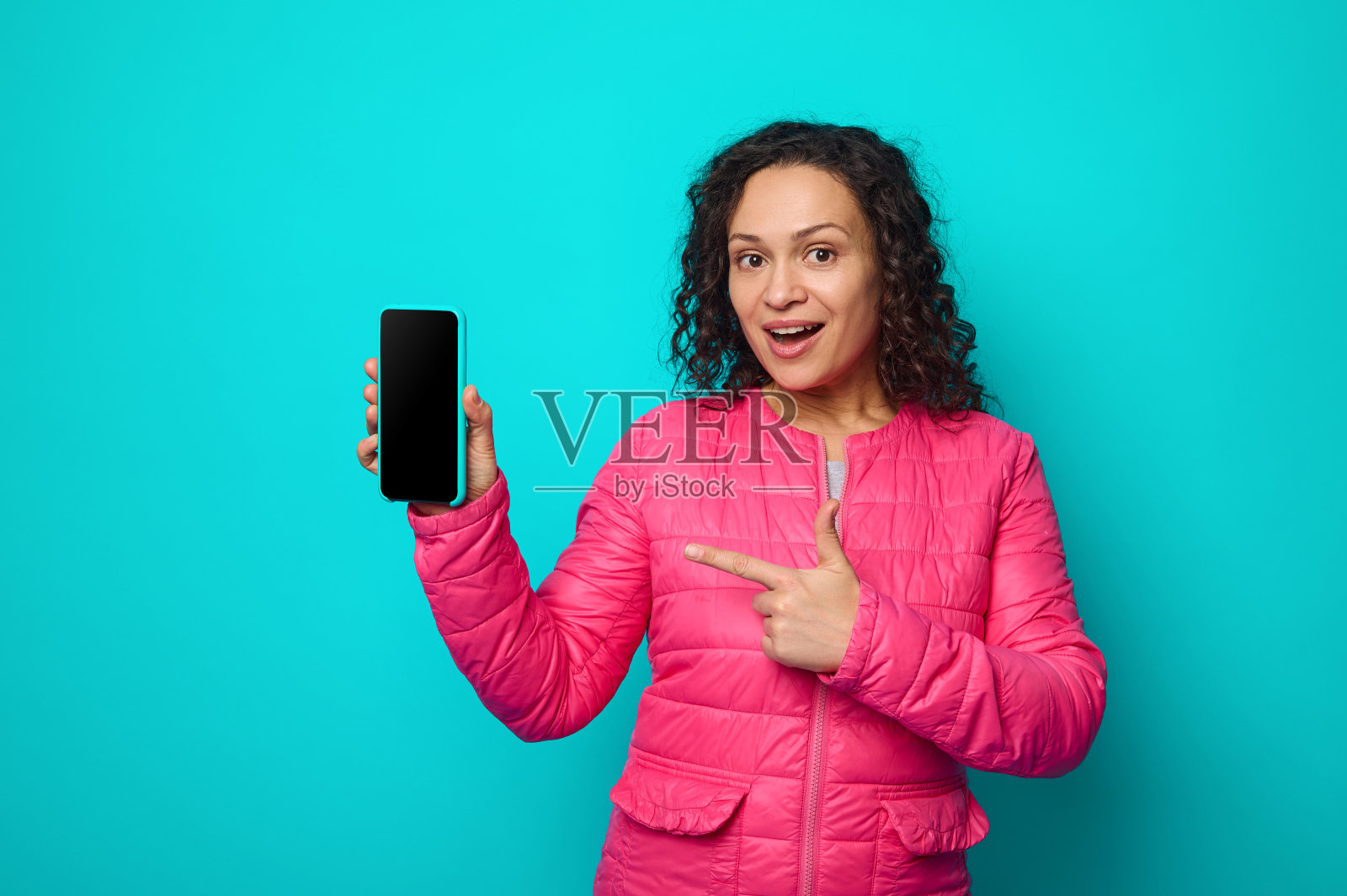 开朗、令人生畏的卷发、穿着亮粉色夹克的迷人女子看着镜头，手指在她手中的智能手机的空白屏幕上，在蓝色背景与拷贝空间上孤立起来照片摄影图片
